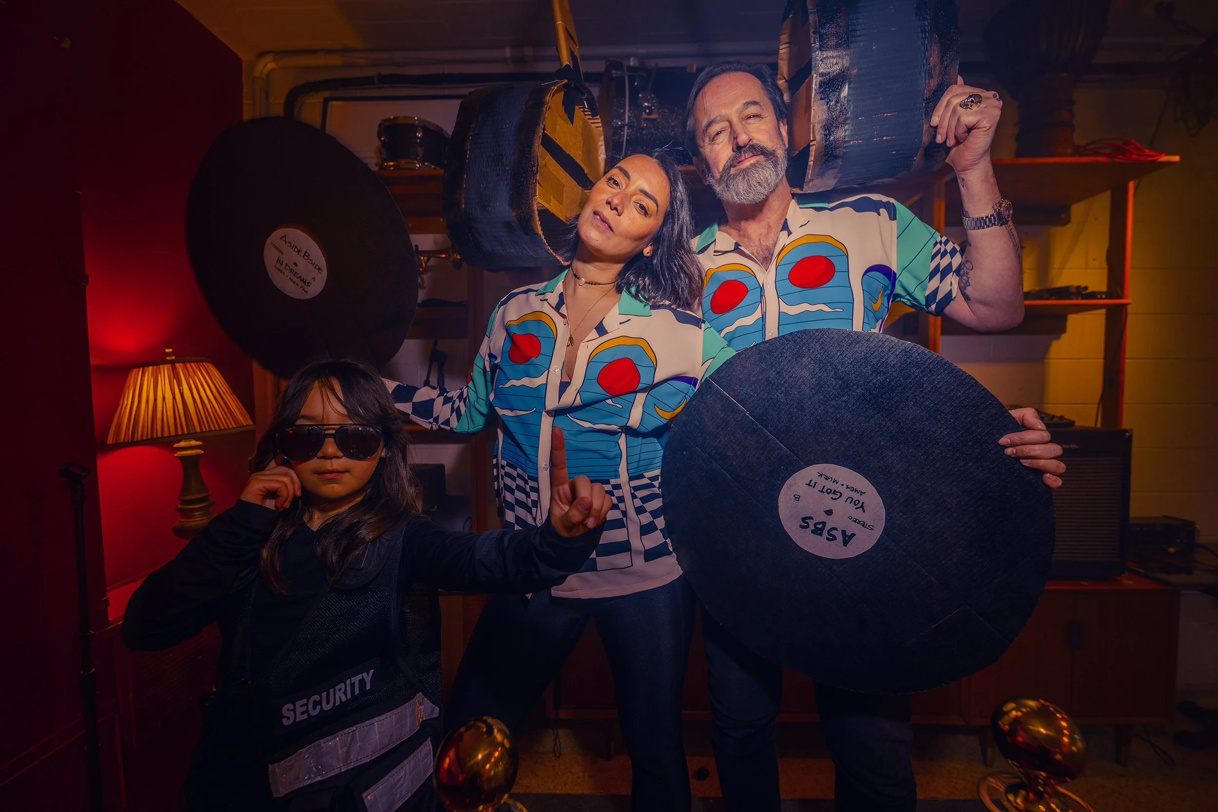A group of four people, including two adults and two children, celebrating in a room with a warm, dimly lit ambiance. They are holding a large vinyl record and making peace signs, wearing colorful, patterned shirts and sunglasses, with Christmas orna