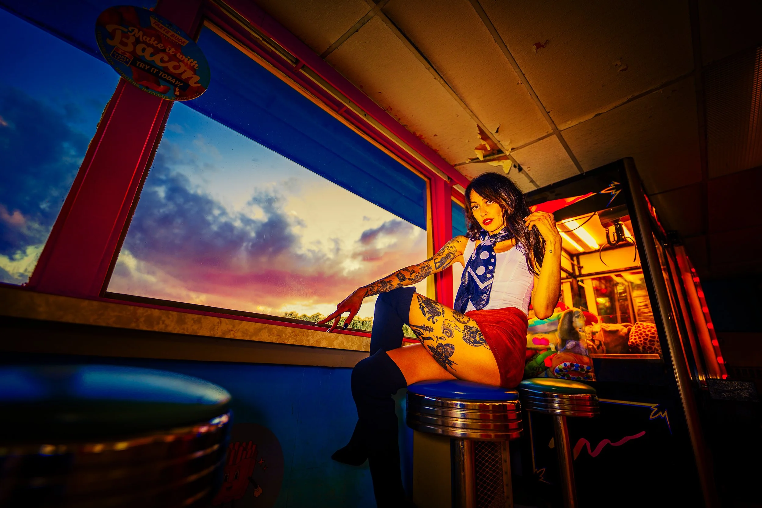 A woman with tattoos and dark hair sits on a barstool inside an arcade or entertainment venue during sunset. She is wearing a white top, red shorts, thigh-high socks, and a polka dot scarf. There are colorful gaming machines and a sunset view through