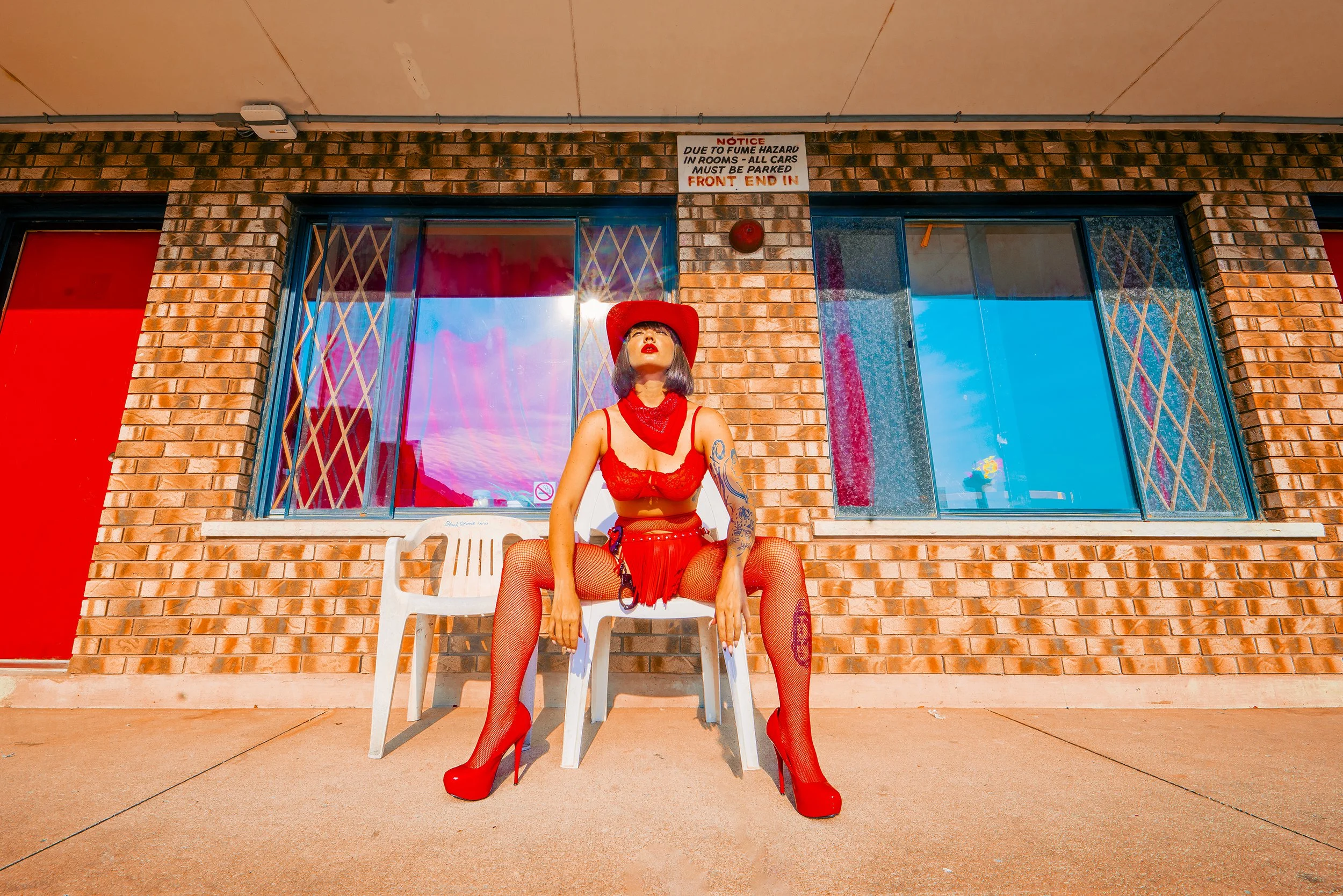 A woman dressed in red with fishnet stockings and high heels sitting on a white plastic chair outside a brick building. She is wearing a red cowboy hat, red bandana, and red lingerie with tattoos on her arm. The sun is shining brightly, reflecting of