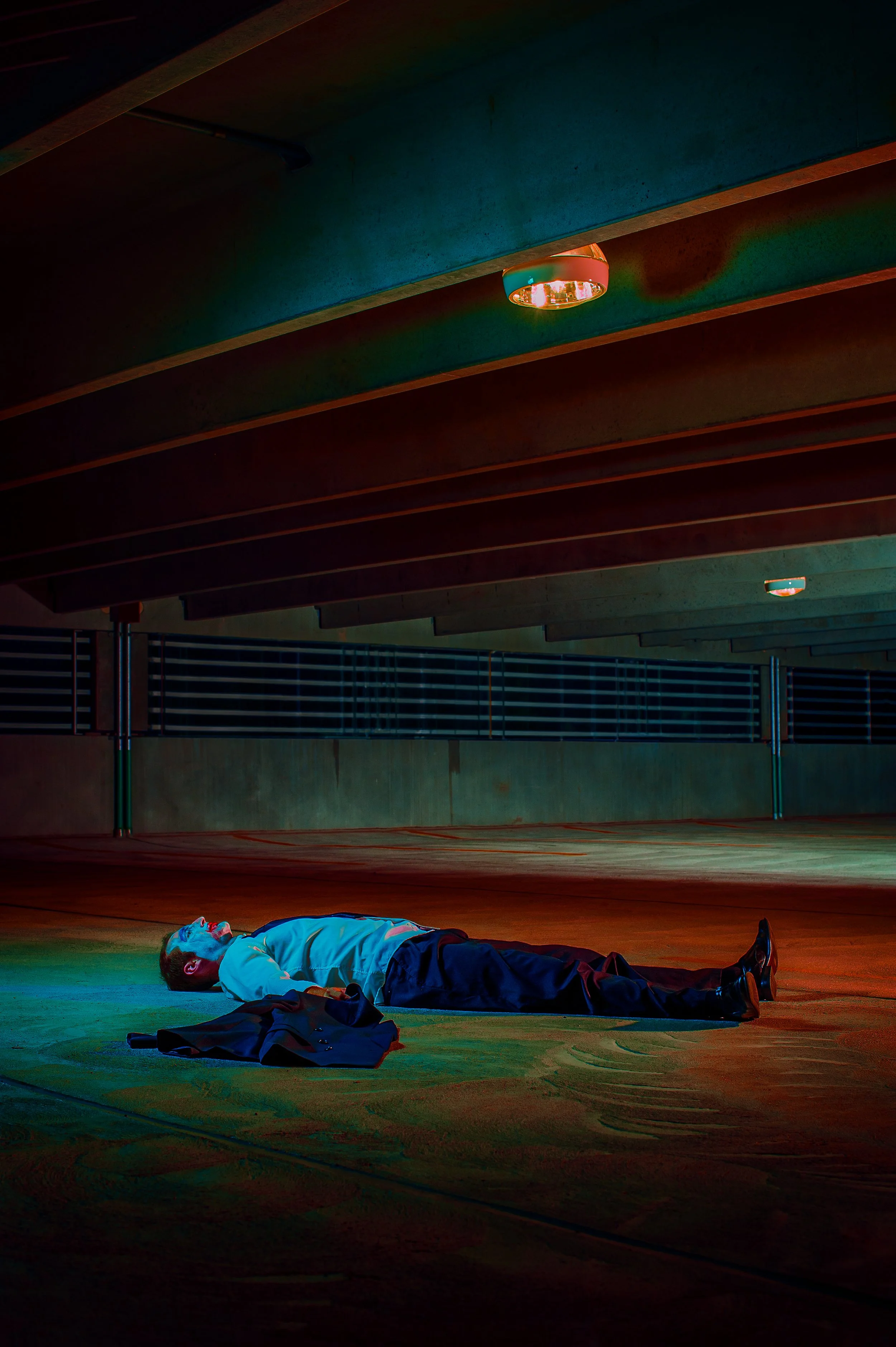 A person lies on the floor of an indoor sports court, possibly resting or unconscious, with a jacket and belongings nearby. The setting features modern lighting and an architectural ceiling with wooden beams.