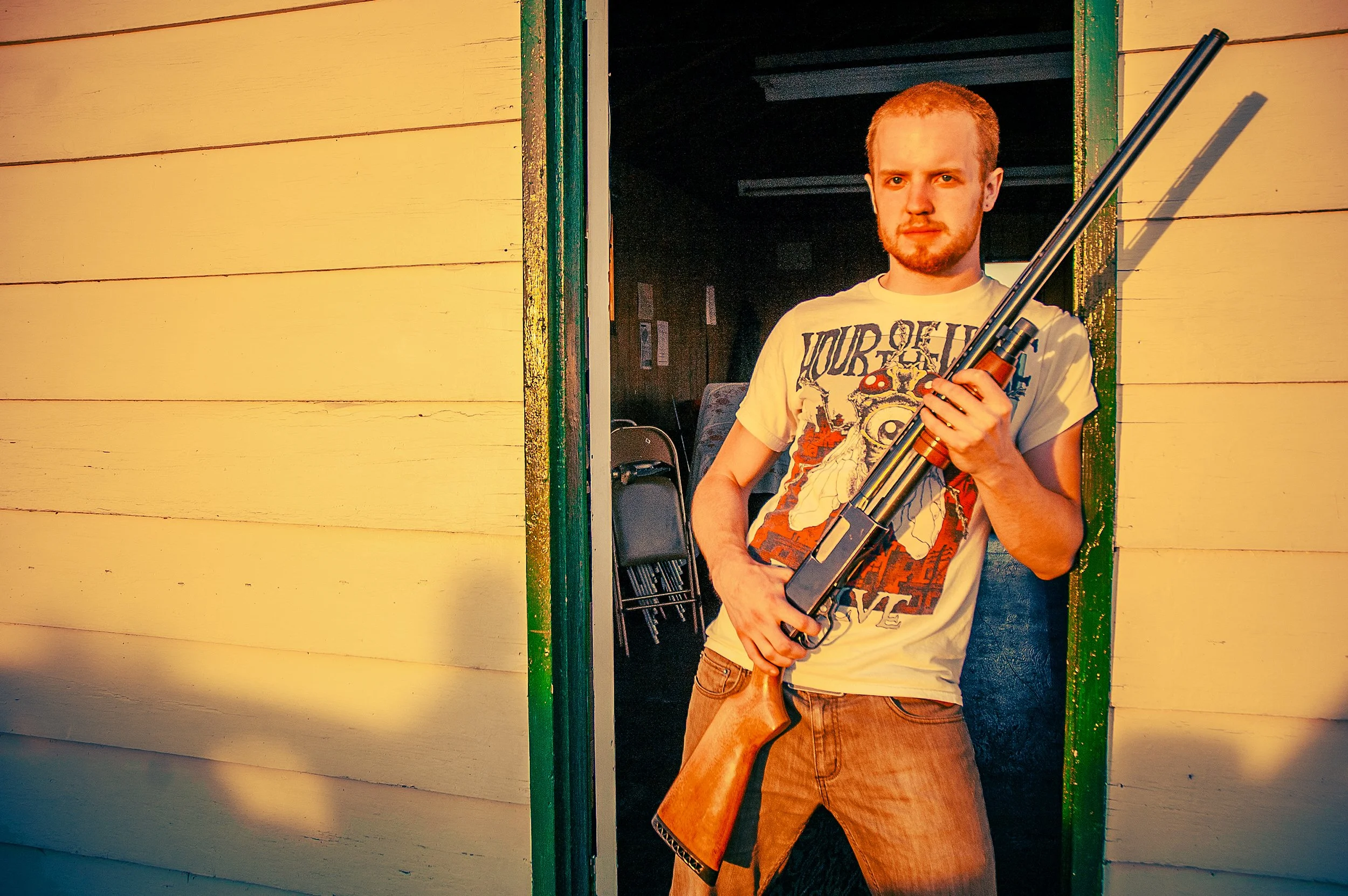 A man with a beard and short red hair, standing in a doorway, holding a rifle, wearing a graphic t-shirt and shorts, with warm sunlight casting an orange glow on the scene.
