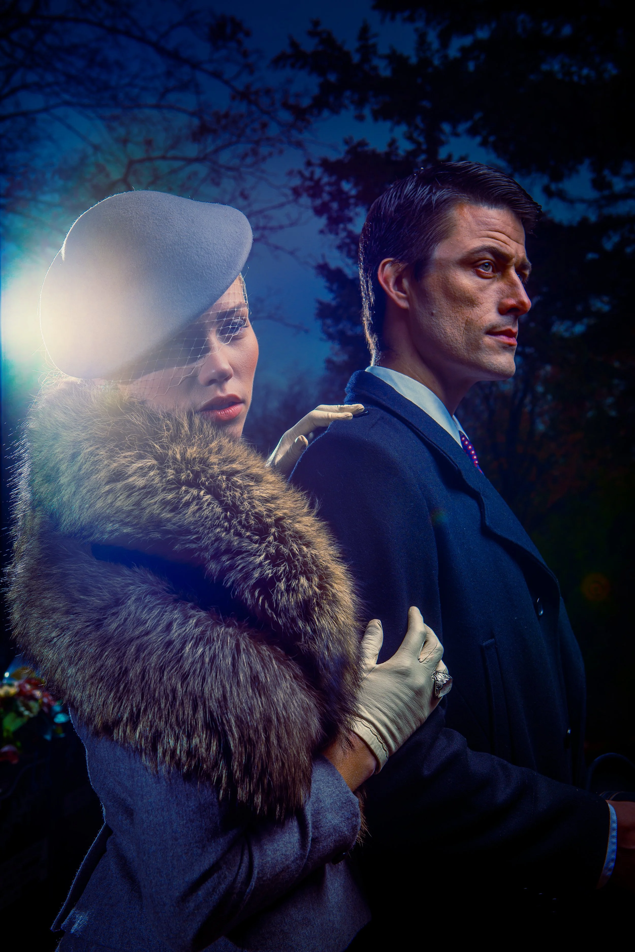 A woman dressed in vintage style clothing with a fur stole and a hat with a veil and netting, standing next to a man in a dark suit. The woman has red lipstick and the man has slicked-back hair, both looking off into the distance during dusk or dawn 