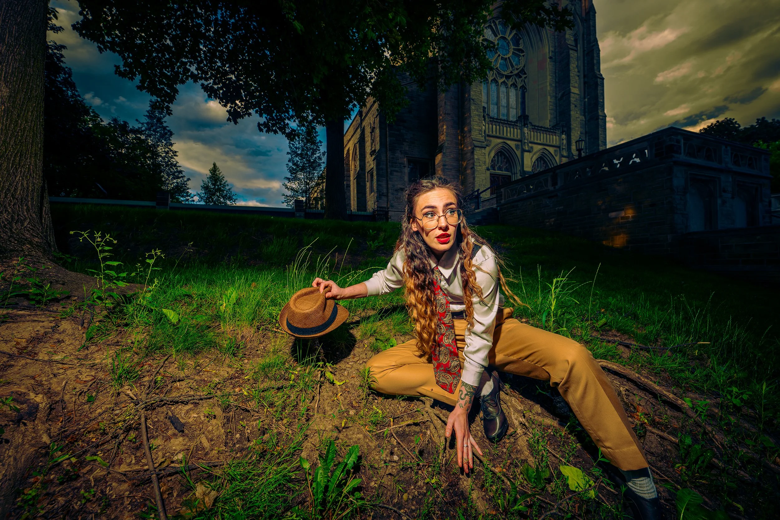 A woman with long curly hair, glasses, and red lipstick lying on the ground in front of a large gothic-style church, holding a tan hat, with green grass, trees, and cloudy sky in the background.