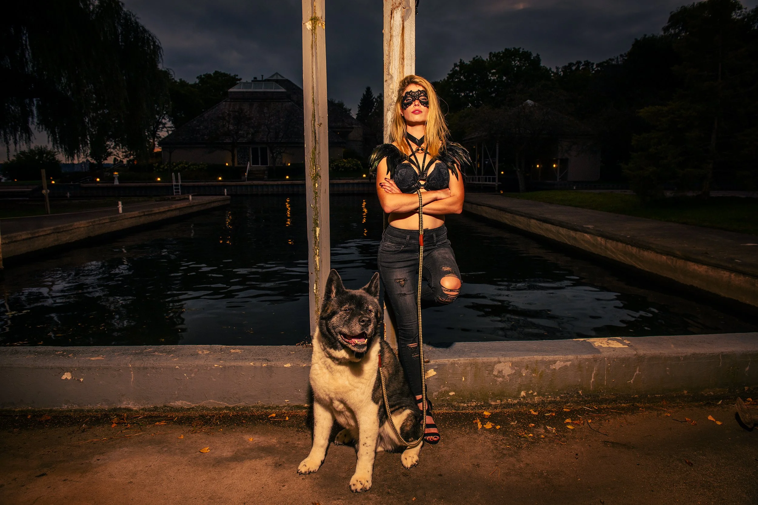 A woman wearing a black lace mask, black top with feather details, and ripped black jeans stands with her arms crossed next to a large black and white dog on a leash, by a canal at dusk.