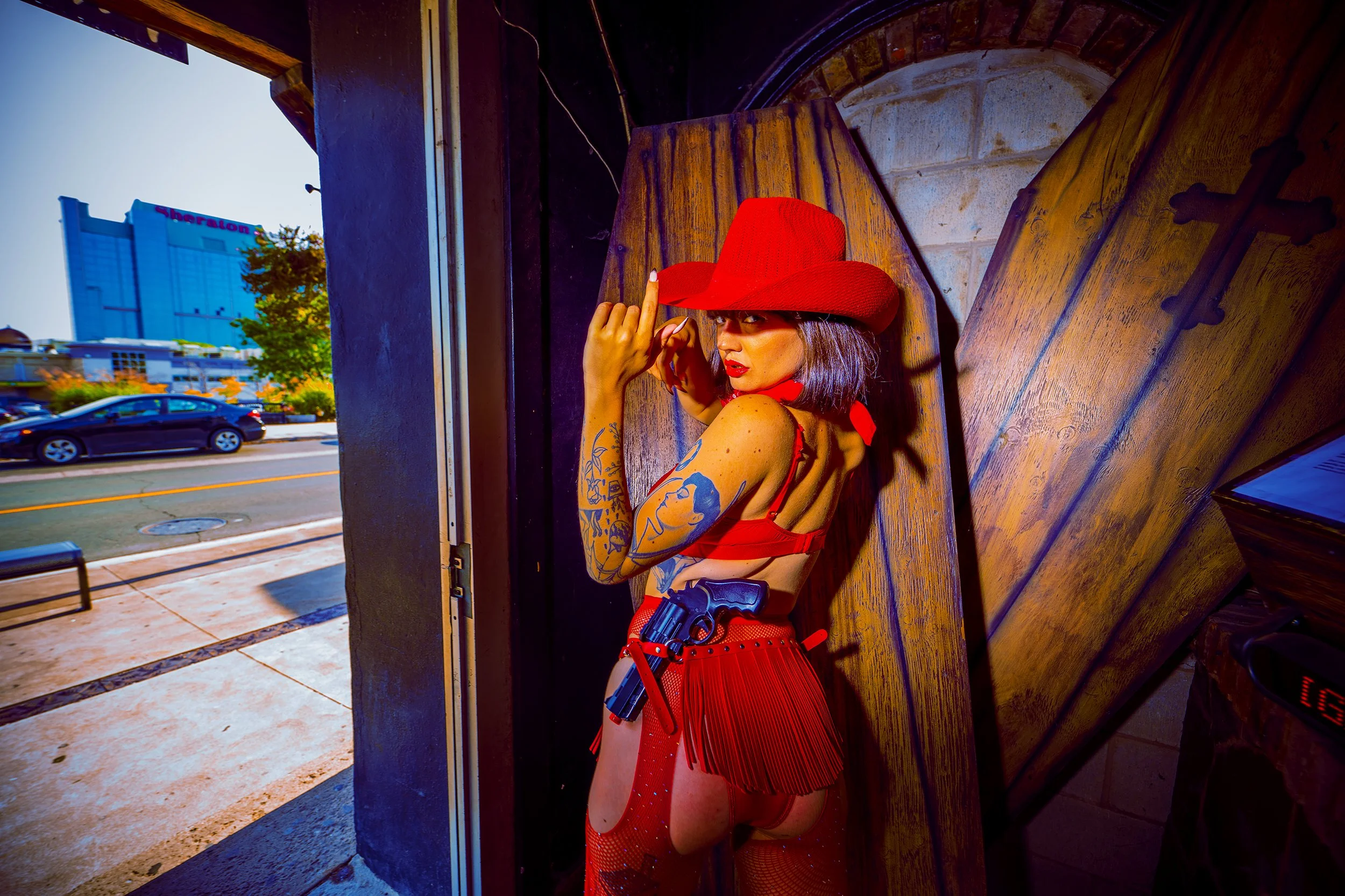 A woman with tattoos wearing a red hat and red outfit, holding a toy gun, posing inside a building with a view of a city street outside.