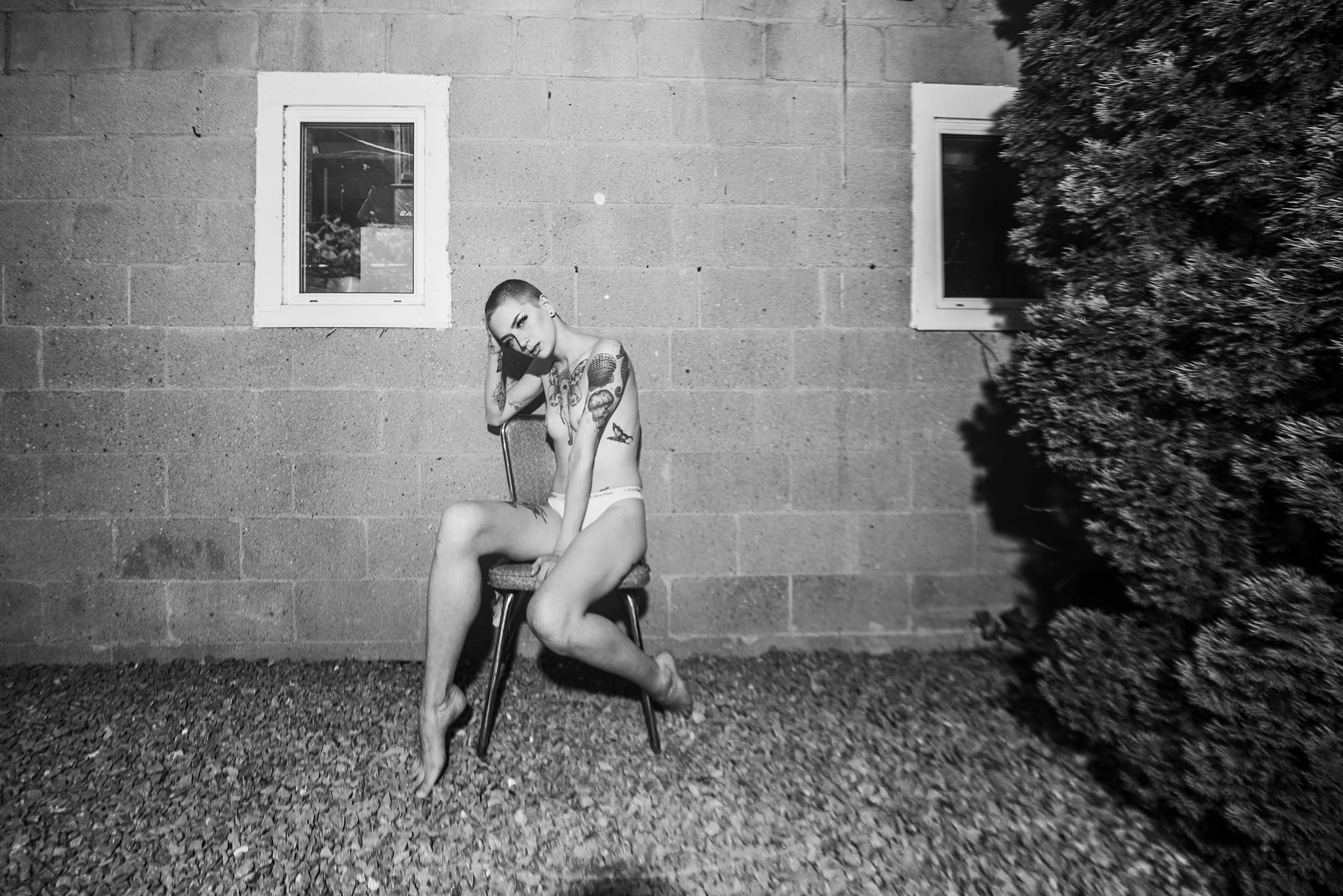 A tattooed woman in underwear sitting on a chair outdoors, with a brick wall and bush in the background, in black and white.