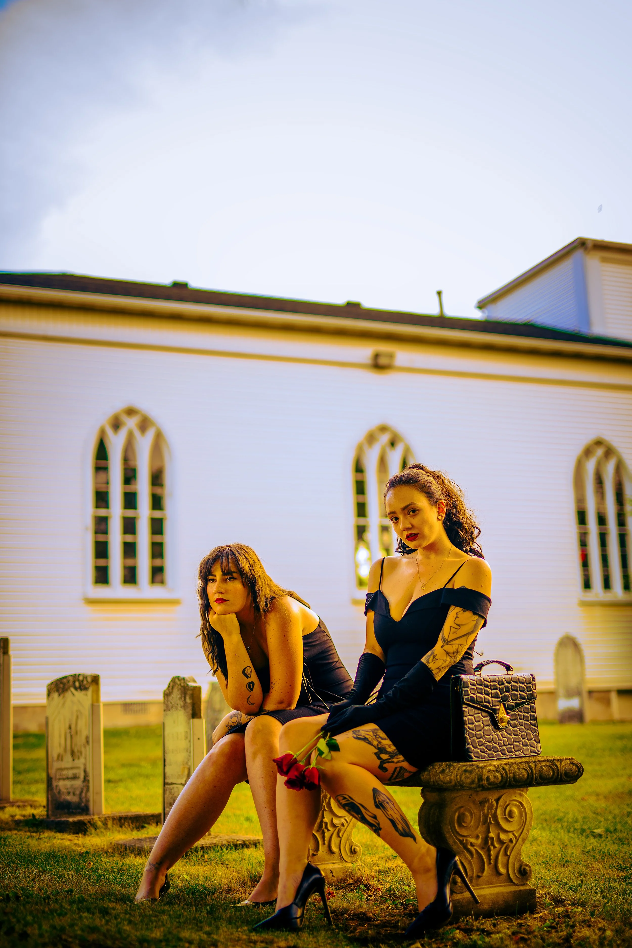 Two women in black dresses sitting on a carved stone bench in front of a white church with arched windows, one holding a red rose, in a grassy area under a cloudy sky.
