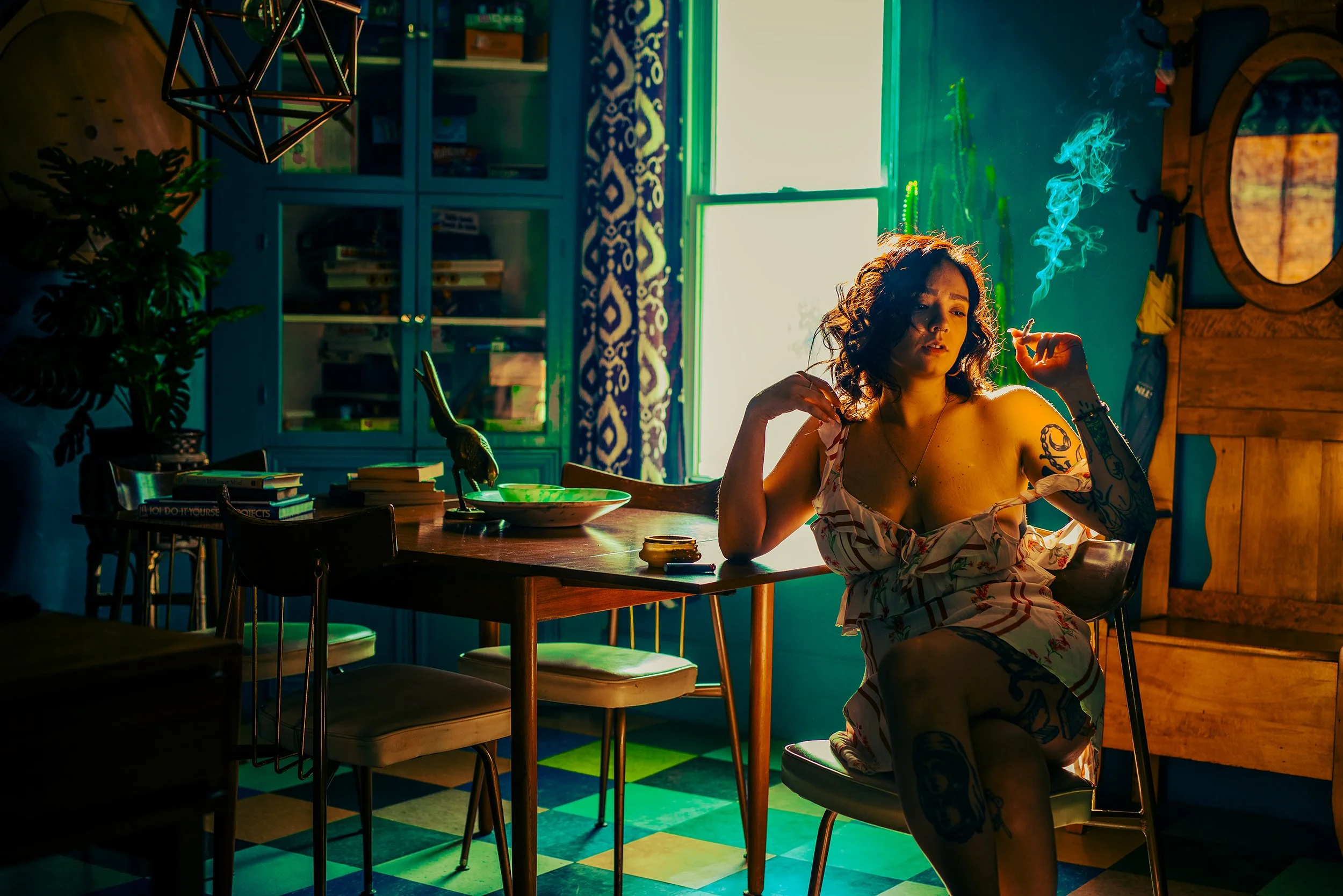 A woman with curly hair and tattoos on her arm is sitting at a table smoking a cigarette in a colorful, dimly lit room with green and yellow light coming from a window.