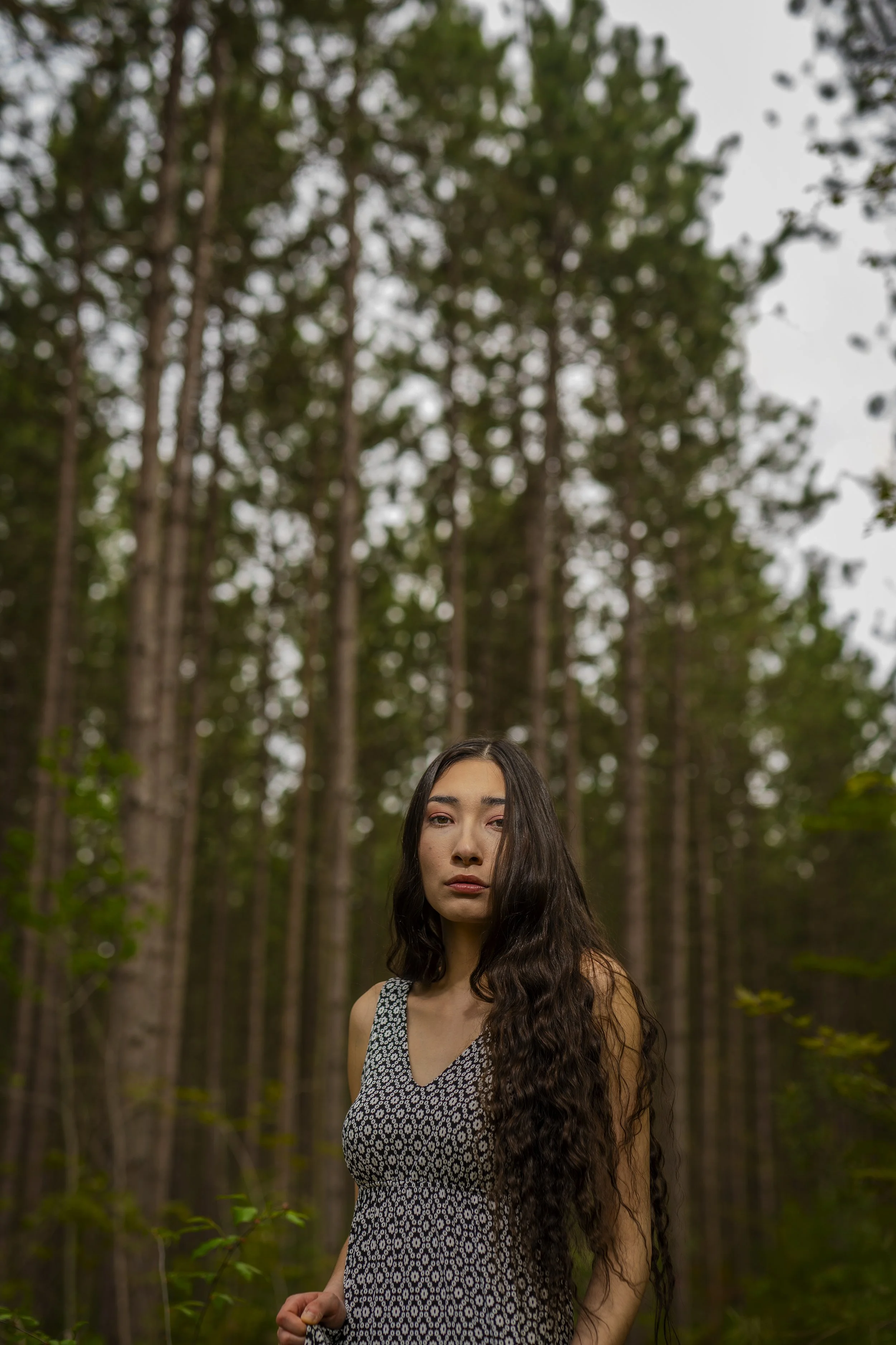 A young woman with long dark hair standing outdoors in a forest, wearing a sleeveless patterned dress, looking at the camera.