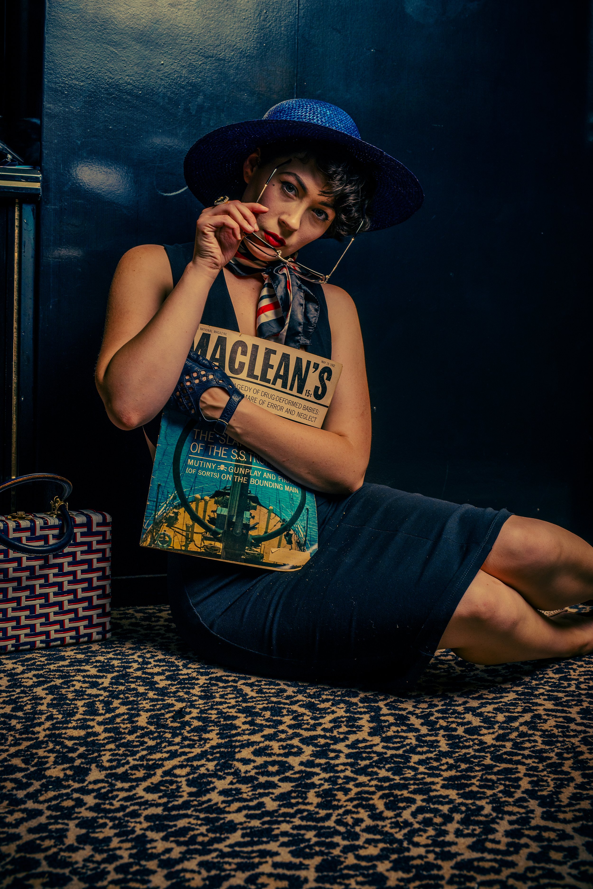 A woman sitting on a patterned carpet, wearing a large blue sun hat, red lipstick, and holding a pair of eyeglasses near her face. She has a vintage-style magazine tucked under her arm, and a patterned bag next to her.