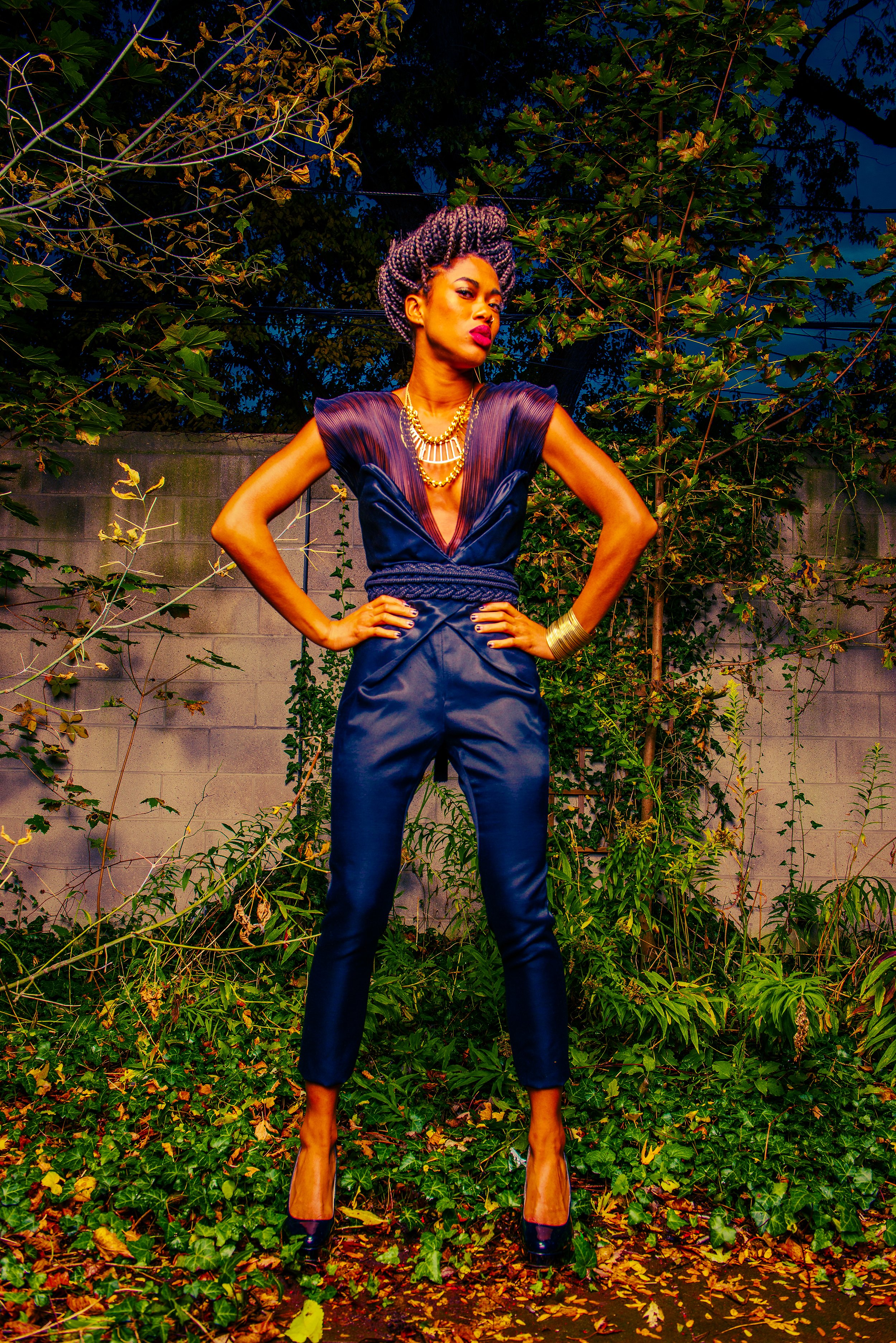A confident woman stands with hands on hips in a dark blue satin outfit and high heels, outdoors at night, surrounded by green foliage and a concrete wall, under a deep blue sky.