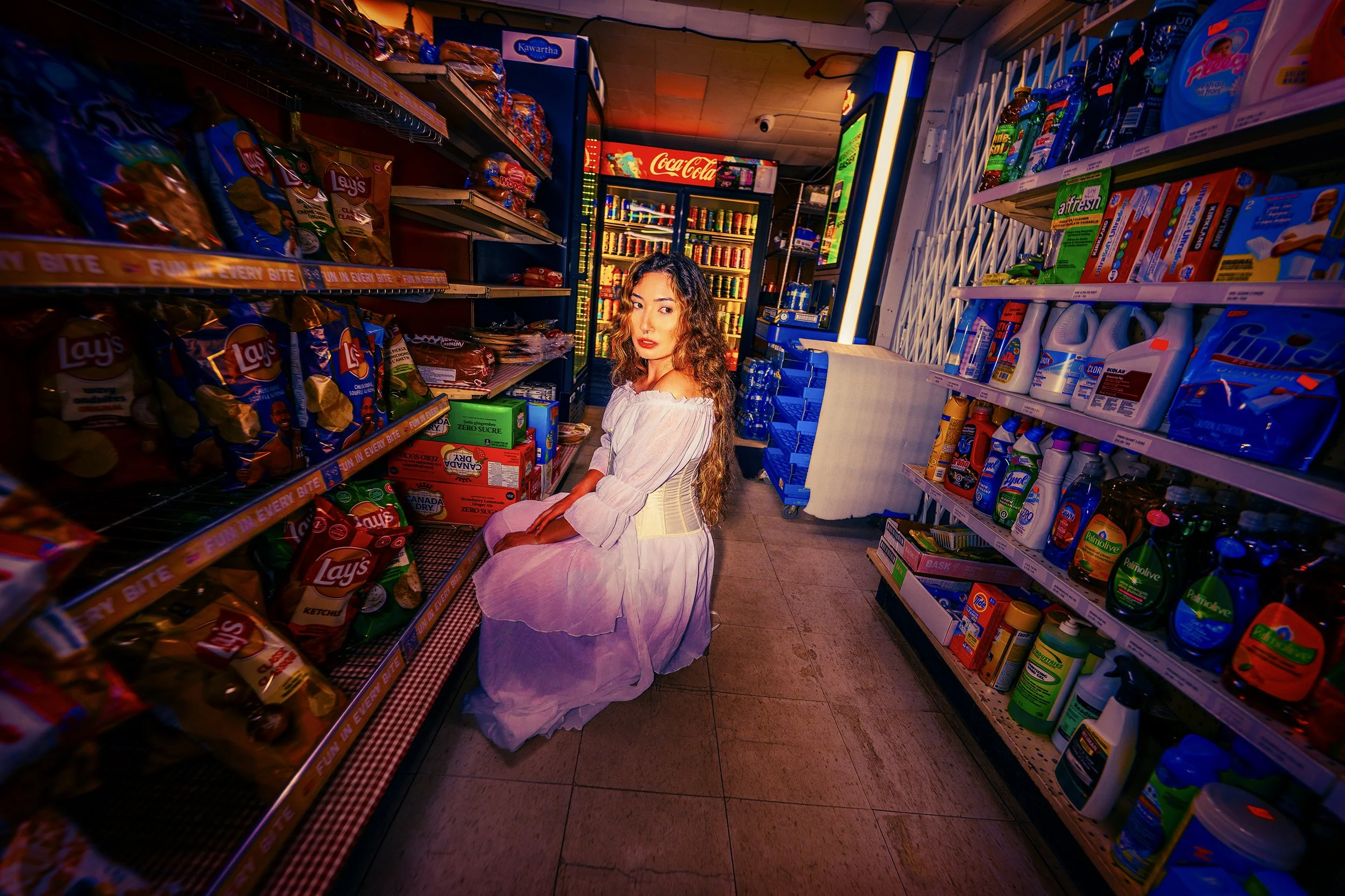 A young woman with long, curly hair sitting on a bench in a grocery store aisle, wearing a white off-shoulder top and a purple skirt, surrounded by shelves stocked with snacks, beverages, and household items.