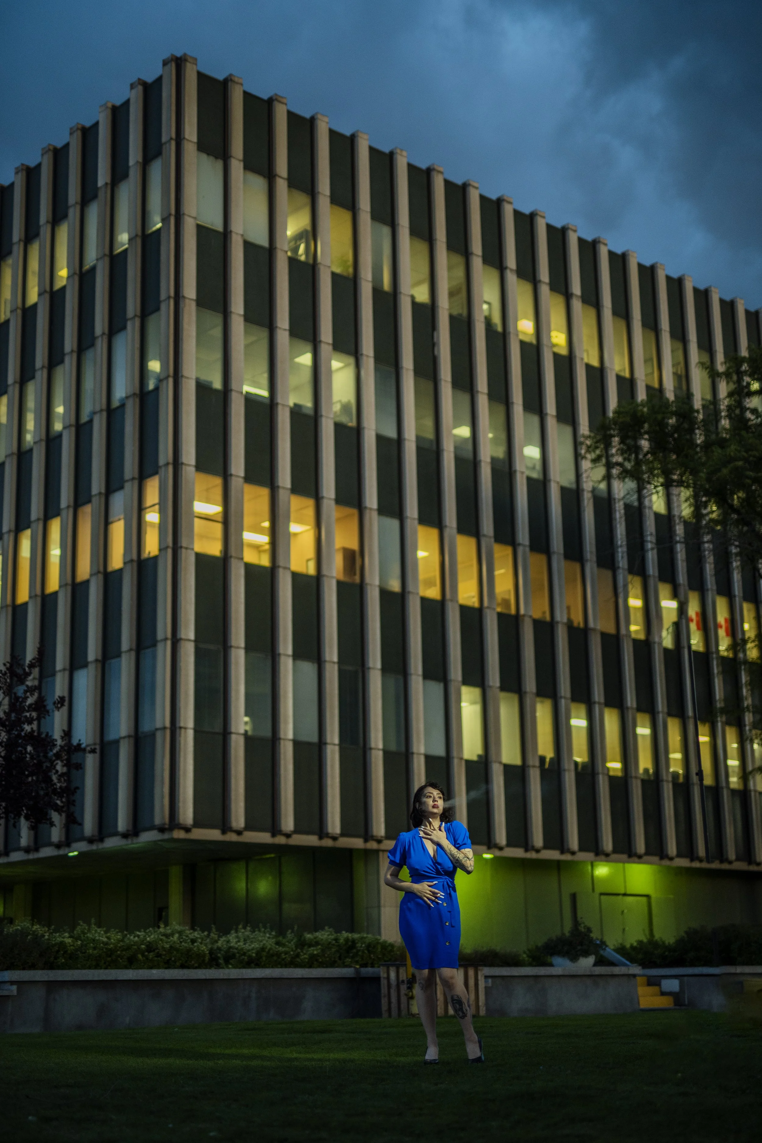 A woman in a blue dress standing outdoors in front of a modern glass building at dusk, with trees and a manicured lawn.