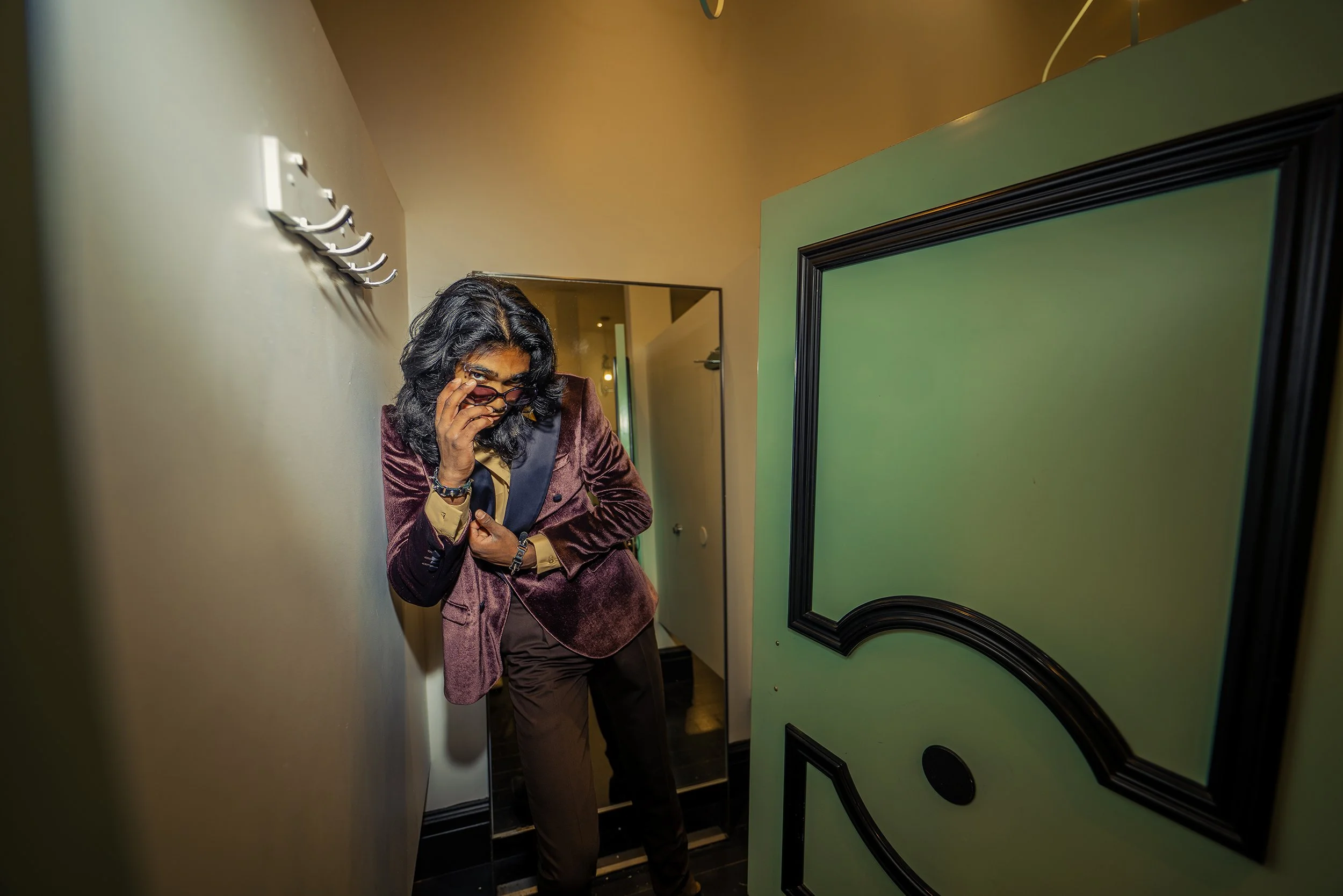 A woman with black, wavy hair and glasses, wearing a velvet jacket, is inspecting her glasses while standing in an elevator. She is looking over her glasses towards the camera.