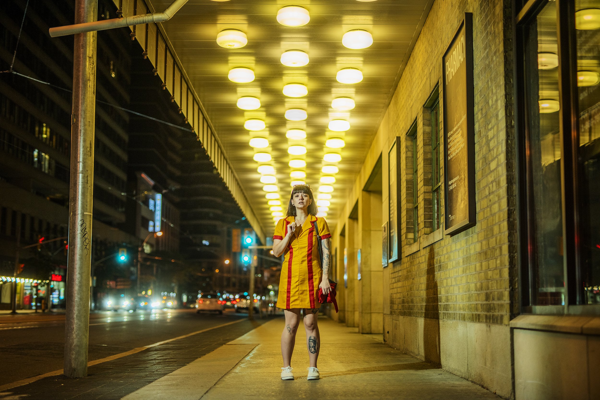 A young woman with tattoos on her legs and arms stands on a city sidewalk at night, wearing a yellow and red striped dress and sneakers, under a yellow-lit ceiling, with traffic lights and buildings in the background.