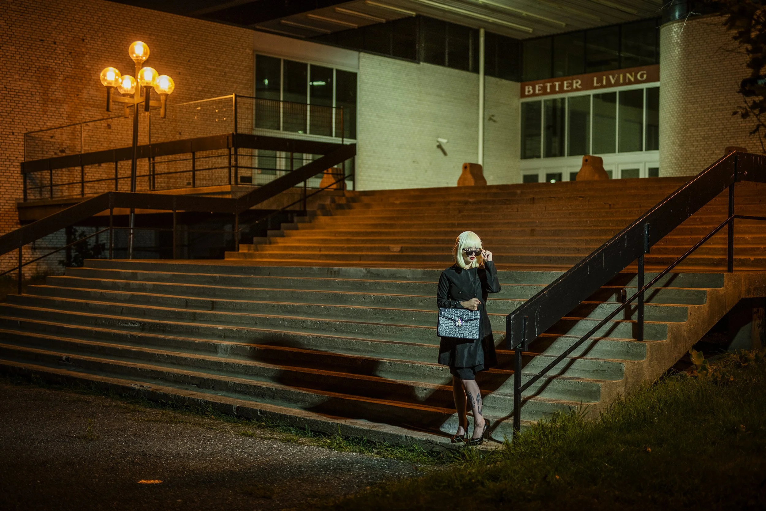 Nighttime scene of a woman with blonde hair, glasses, and a designer handbag, standing on outdoor stairs in front of a modern brick building with large glass windows and a sign reading 'Better Living.' A street lamp illuminates the scene, and there a