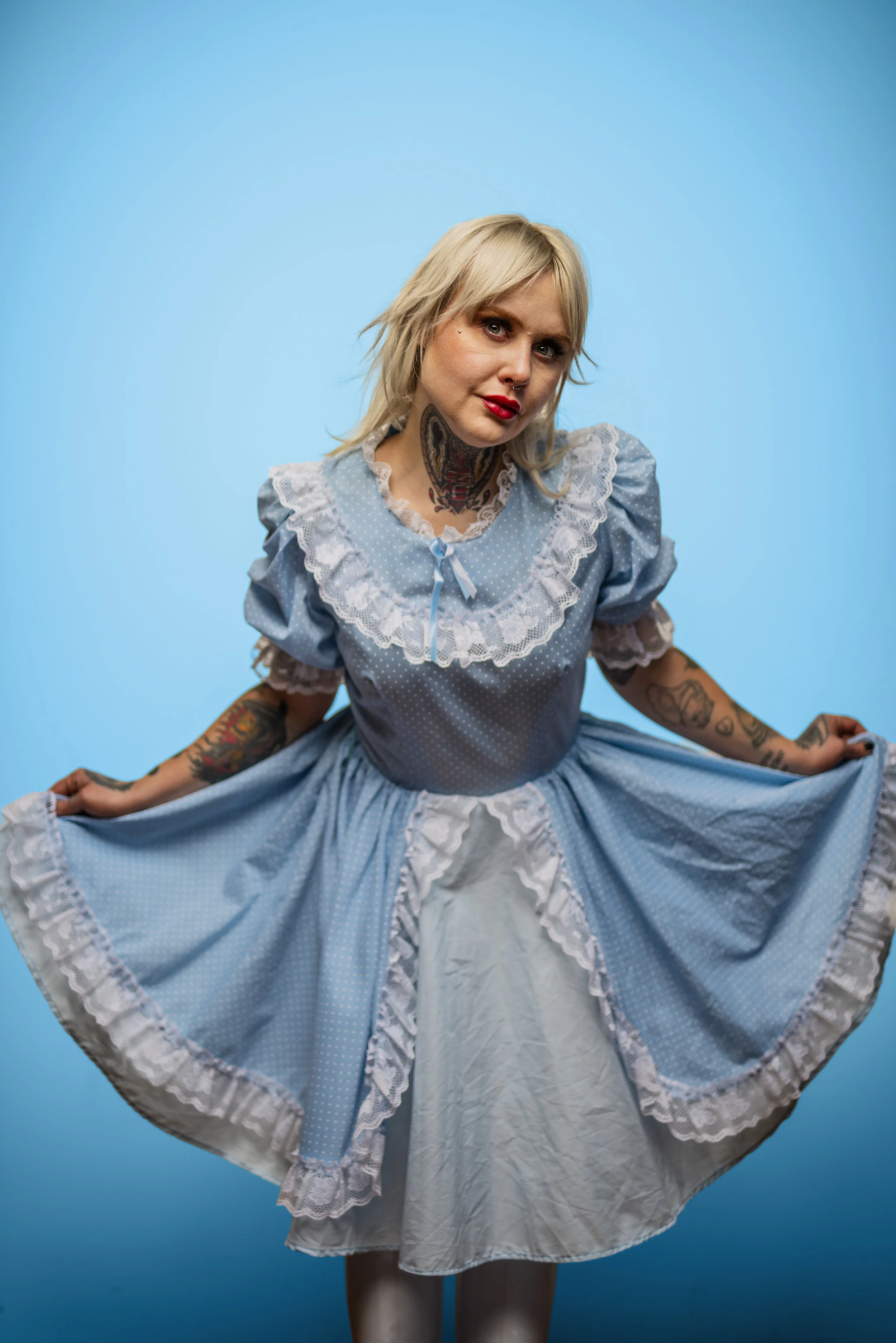 A woman dressed in a vintage blue dress with white lace trim, lifting the skirt to reveal white petticoats underneath, against a bright blue background.