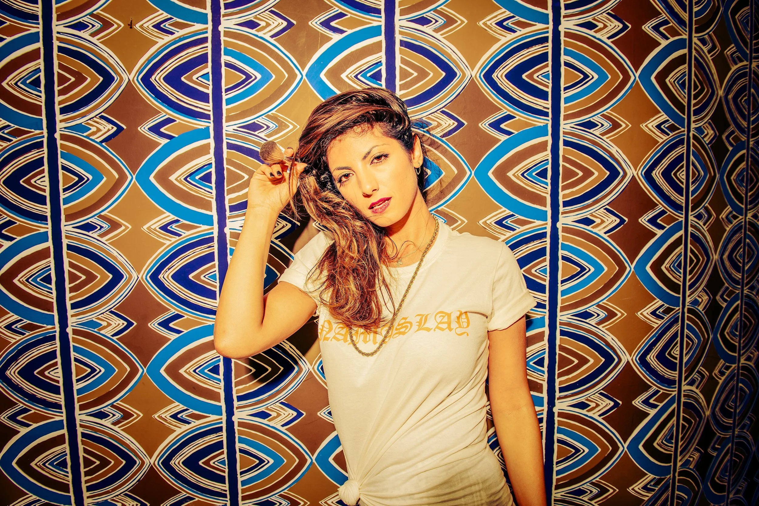 A woman with long wavy hair and light skin holding a makeup brush next to her face, standing in front of a colorful geometric patterned background.