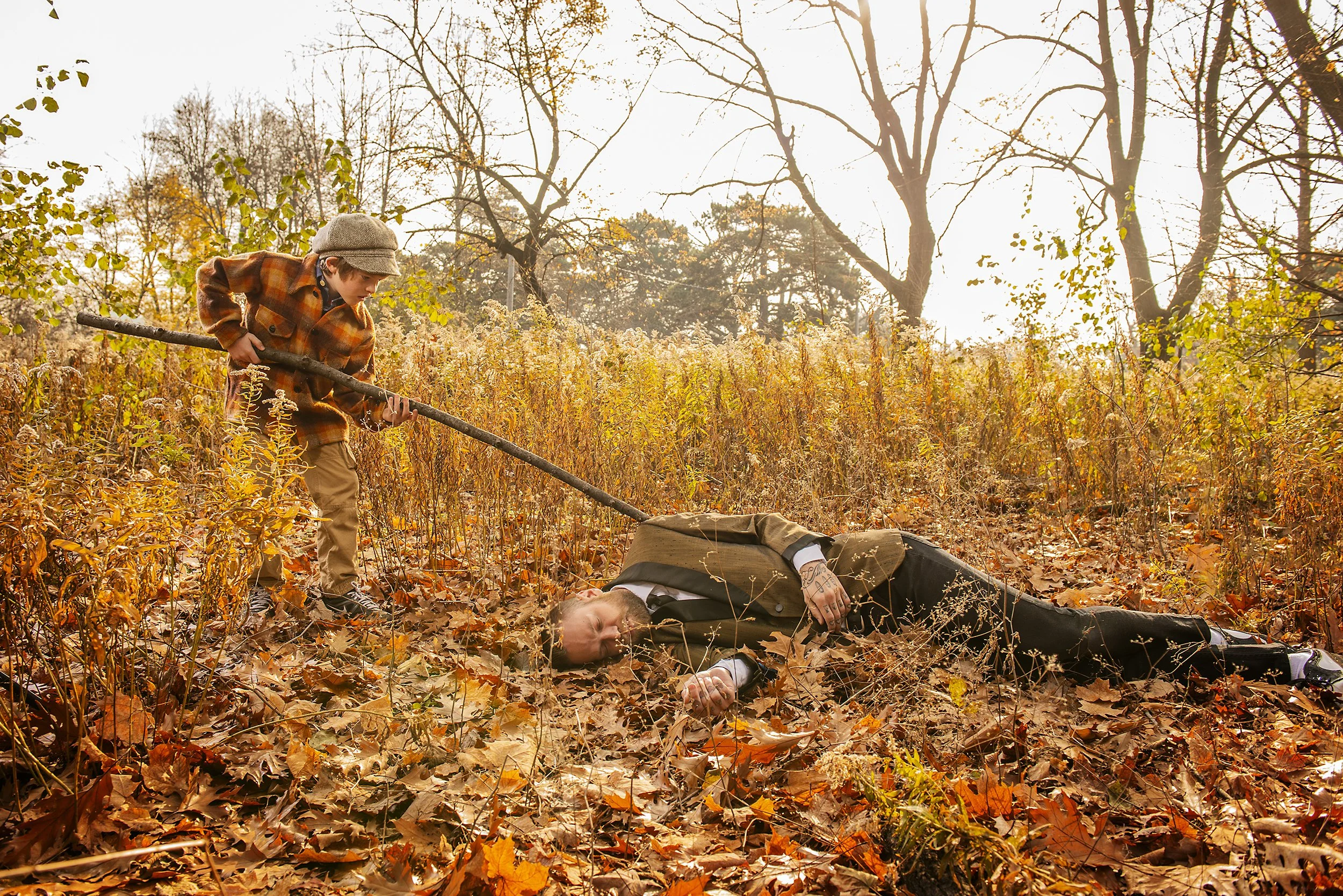 A person lying on the ground dead with fallen leaves in a park during autumn, another person standing nearby holding a stick, wearing a plaid coat and hat, with trees in the background.