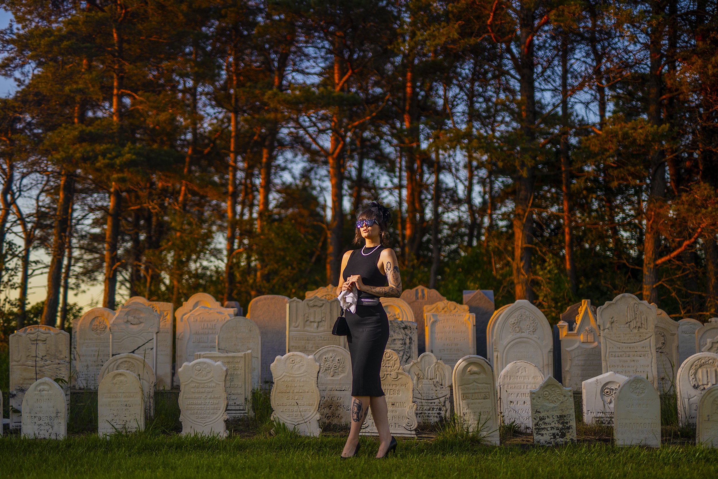 A woman dressed in black, wearing sunglasses and high heels, standing among rows of gravestones in a cemetery at sunset, with tall trees in the background.