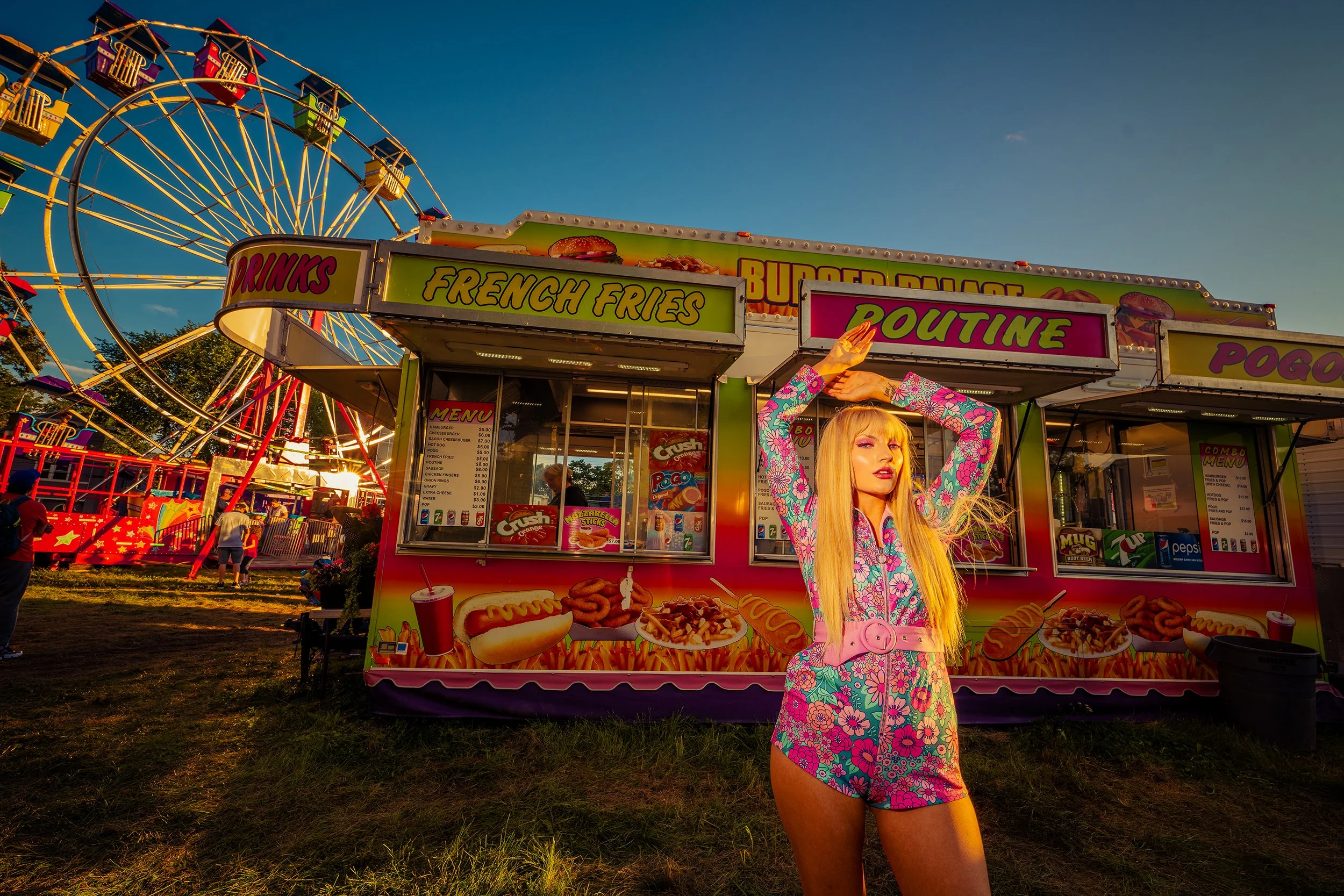 A woman with long blonde hair and bright makeup posing in front of a colorful carnival food stand with a Ferris wheel in the background on a sunny day.
