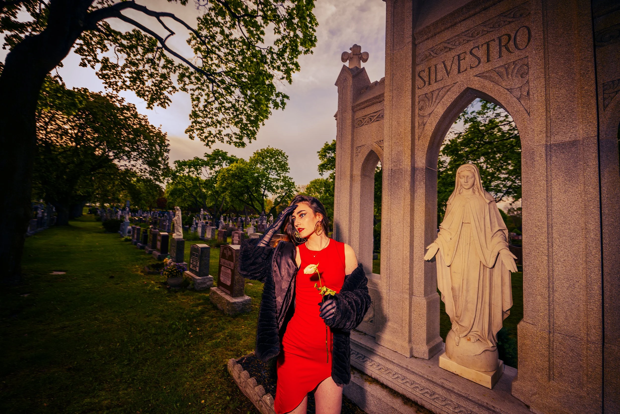 A woman in a red dress with black gloves and earrings stands next to a statue in a cemetery at sunset. She is holding a white flower and has styled hair and makeup.