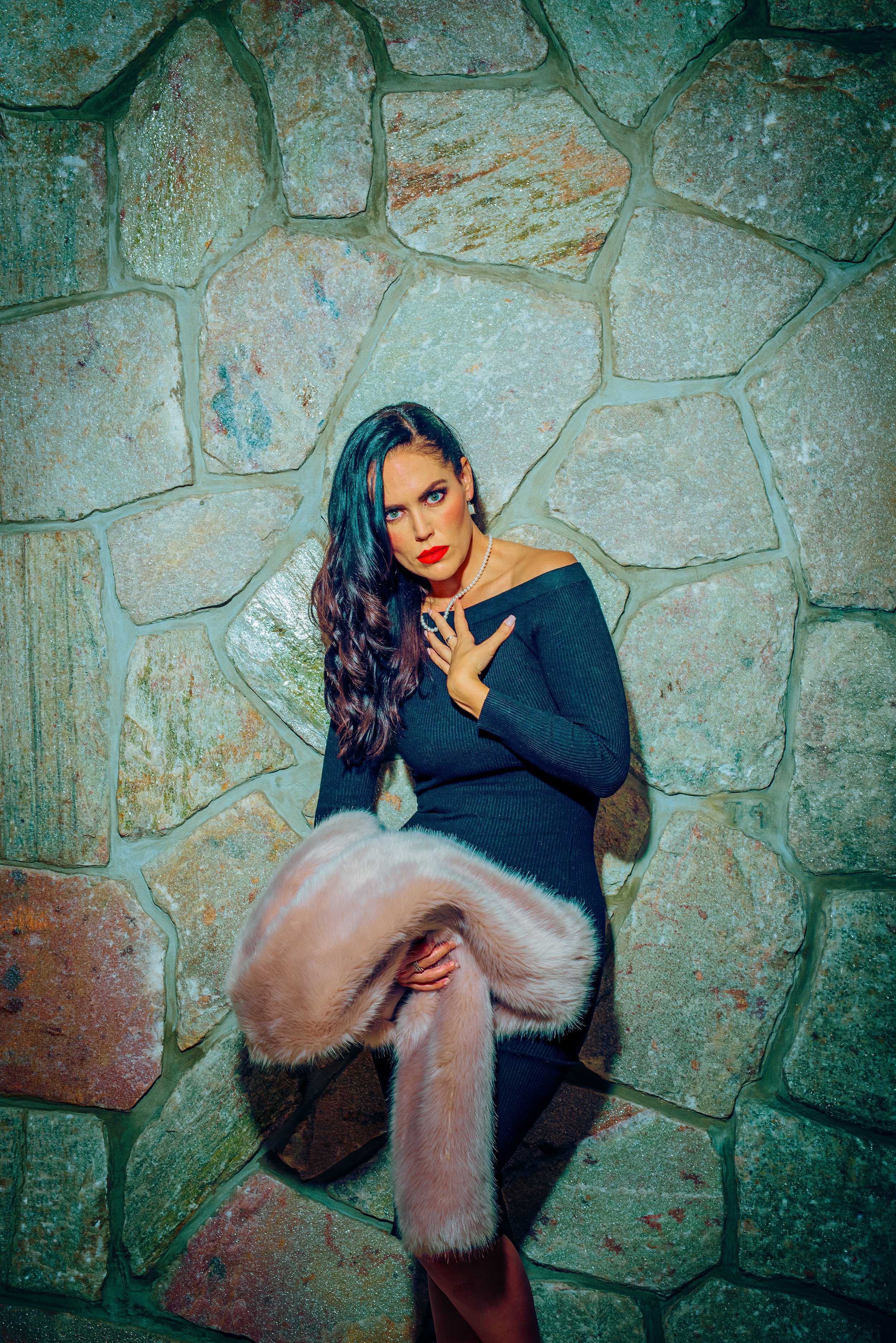 A woman with long dark hair, blue eyes, and red lipstick wearing a black off-the-shoulder top, holding a pink faux fur coat, standing against a large stone wall with uneven, multi-colored stones.