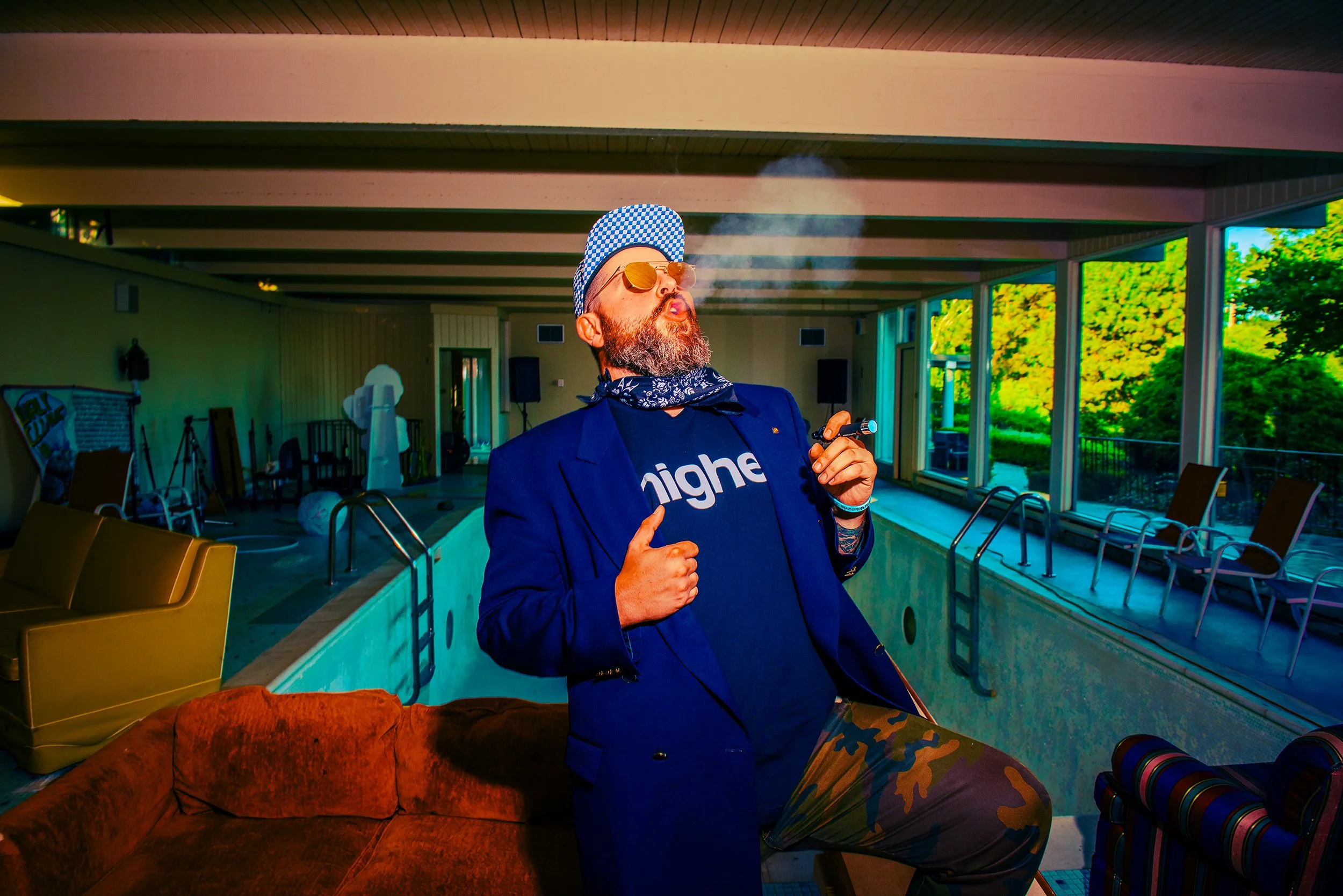 A man with a beard, wearing glasses, a checkered cap, a bandana, a dark blazer, and camouflage pants, is smoking and holding a vaping device indoors near an empty pool area.