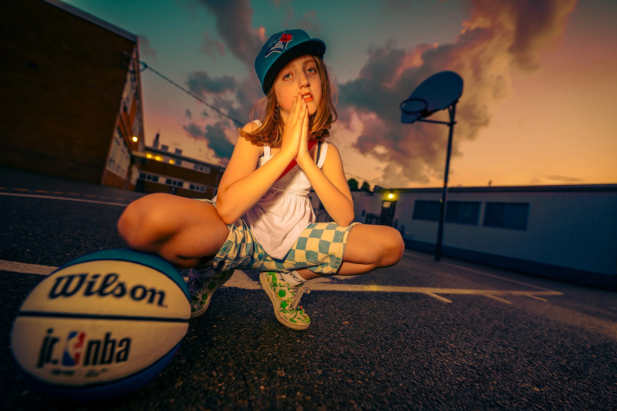 A young girl sitting cross-legged on an outdoor basketball court at sunset, wearing a Toronto Blue Jays hat, decorative sneakers, and plaid shorts, with a Wilson NBA basketball in the foreground.