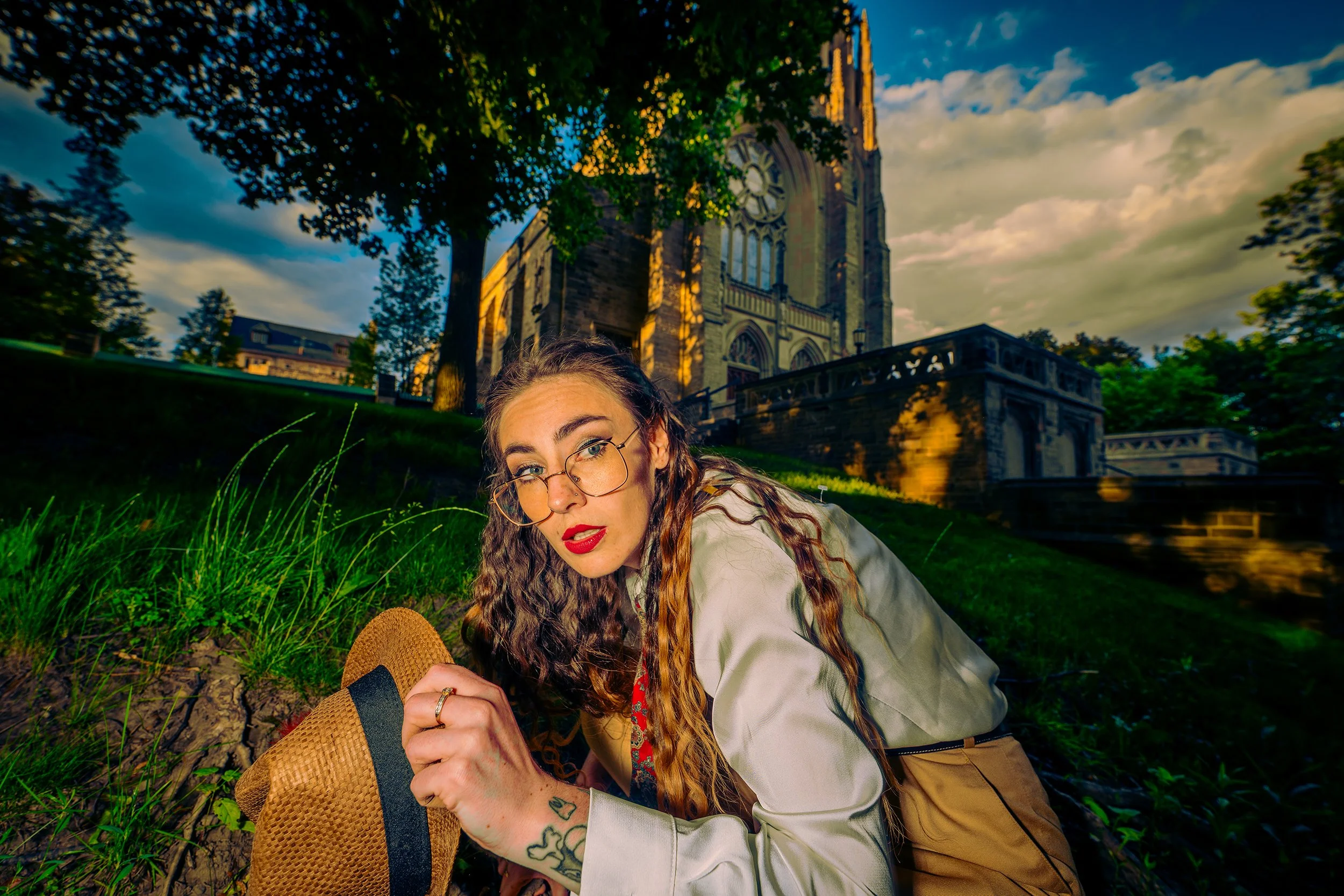 A young woman with long curly hair and glasses lying on the grass in front of a large church with a tall clock tower, surrounded by trees, during sunset.