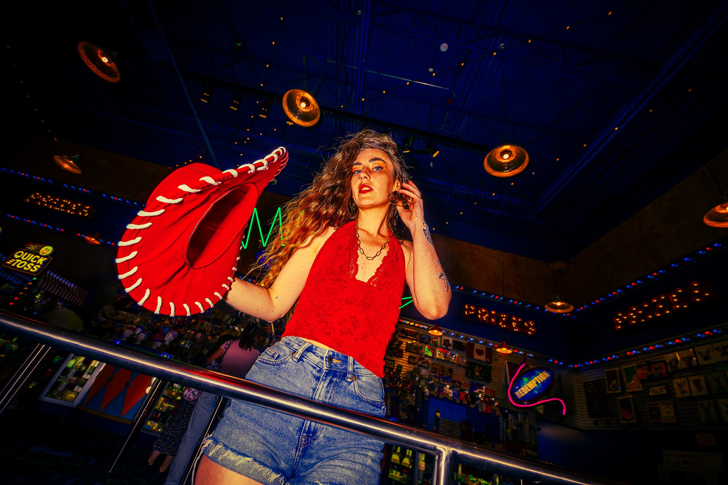 A woman with curly hair wearing a red sleeveless top and denim shorts standing in an arcade. She is holding a red and white plush handbag resembling a baseball glove and talking on a cell phone. The arcade is brightly lit with colorful signs and game