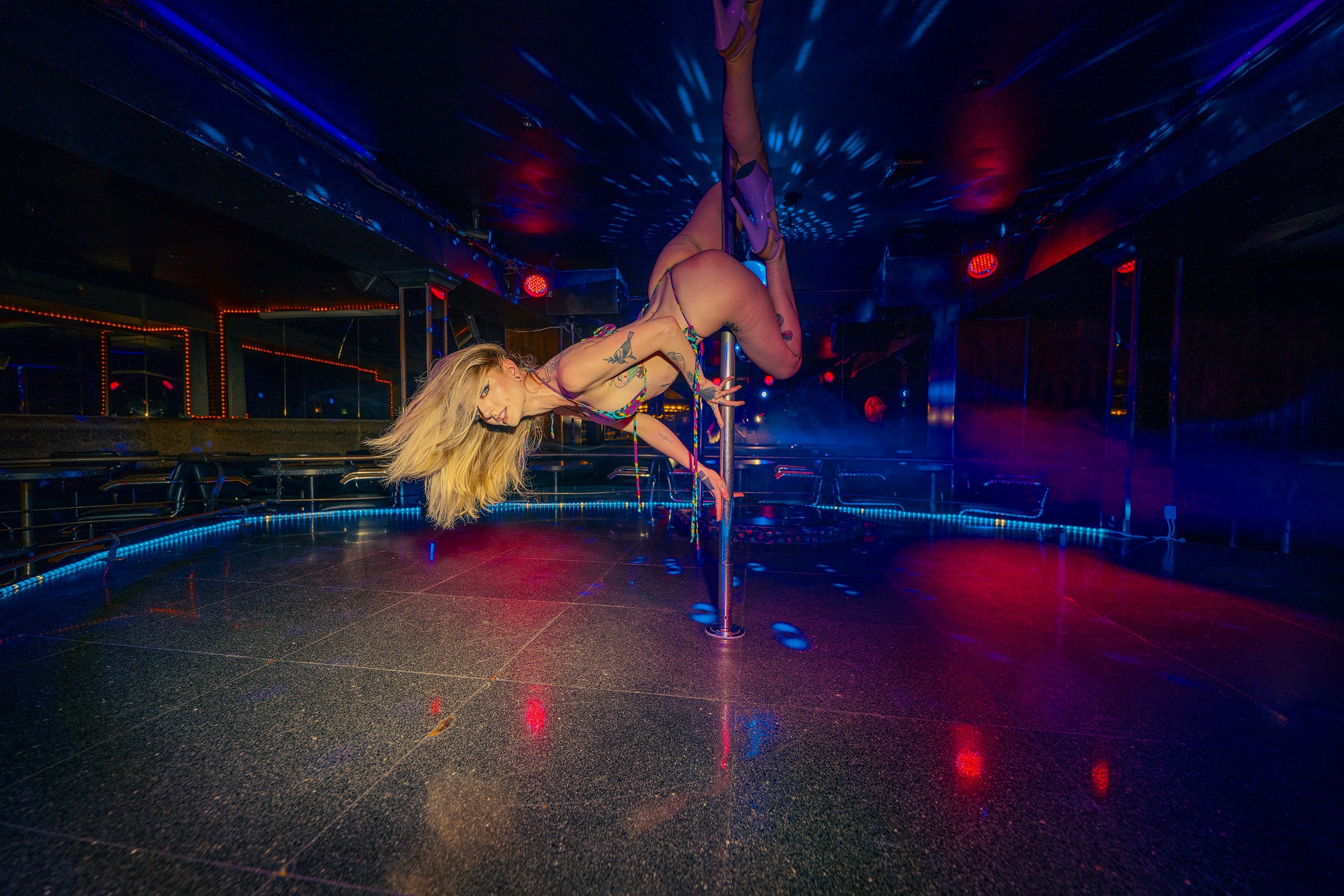 A woman with long blonde hair performs a pole dance in a dimly lit nightclub, surrounded by colorful lights and reflections on the floor.
