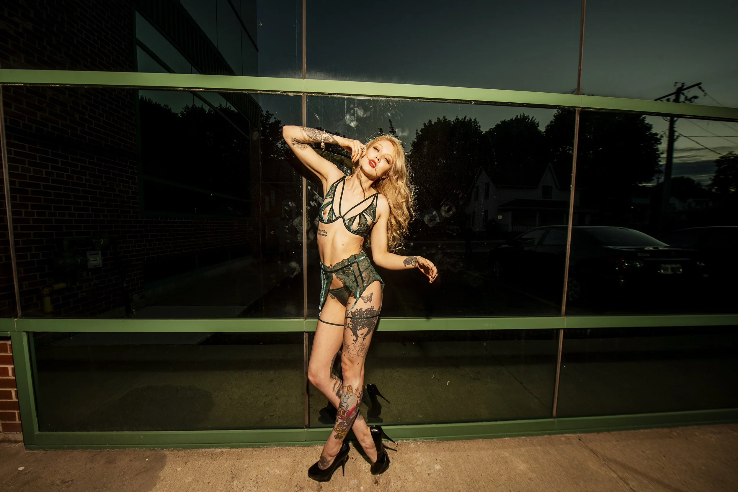 A woman with long blonde curly hair and tattoos on her legs and arms, standing in front of a large reflective glass window, wearing black and green lingerie and high heels, posing with her hand near her face during dusk.
