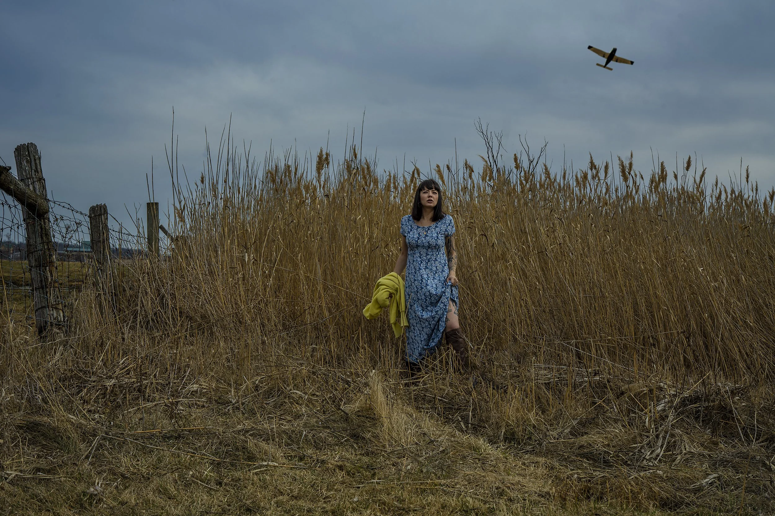 A woman in a blue dress with floral pattern stands in tall, dry grass on a cloudy day, holding a yellow jacket, with a small airplane flying overhead and a weathered wooden fence to the left.