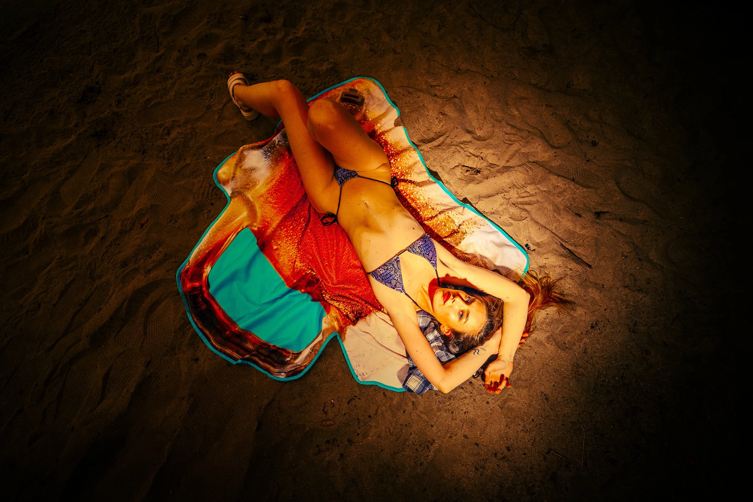A woman lying on a colorful towel on the sandy beach, wearing a bikini, with her arms behind her head and smiling at the camera.