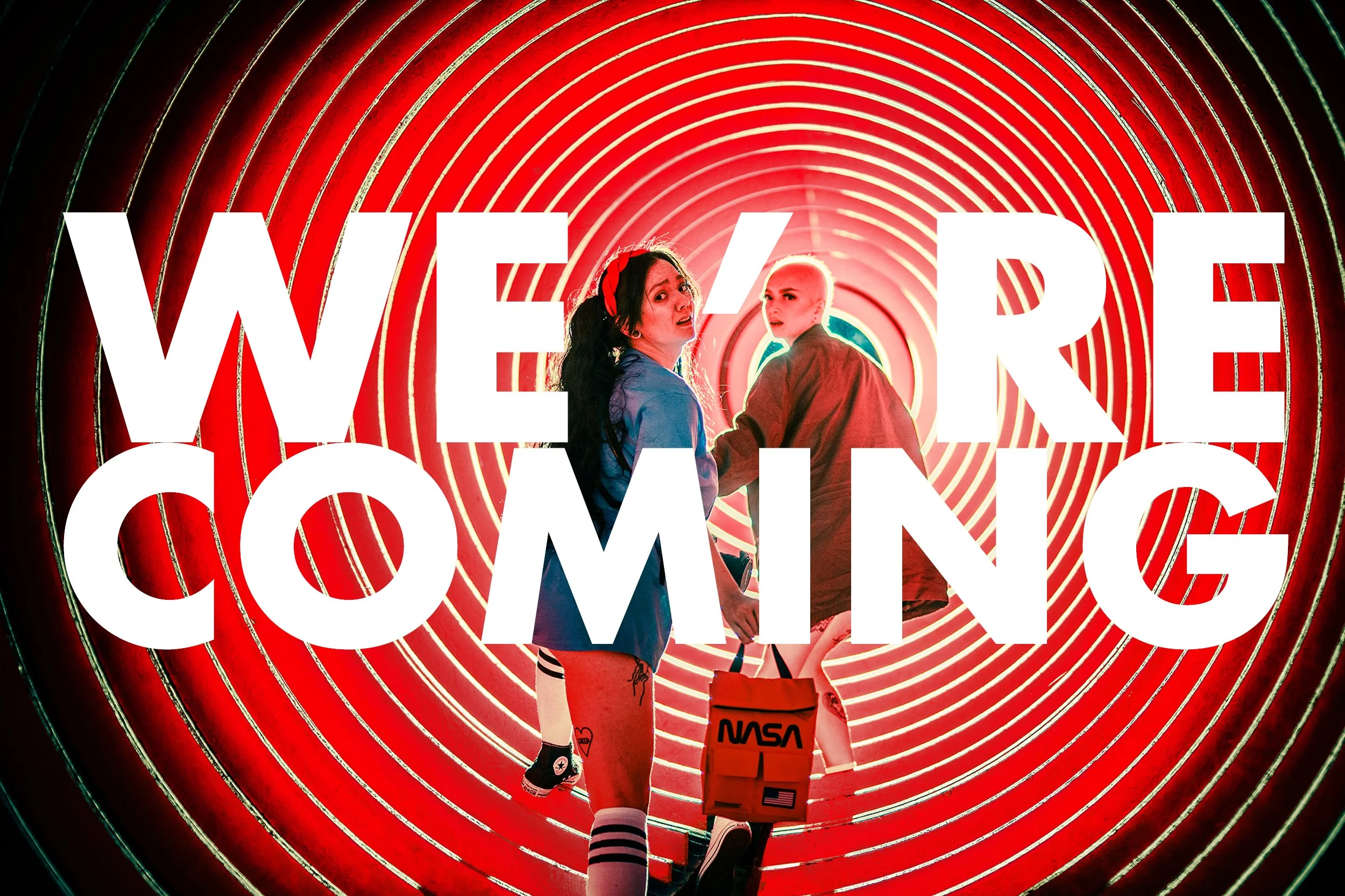 Two women standing in front of a glowing, circular, spiral tunnel with the large text 'WE ARE COMING' overlayed in white.