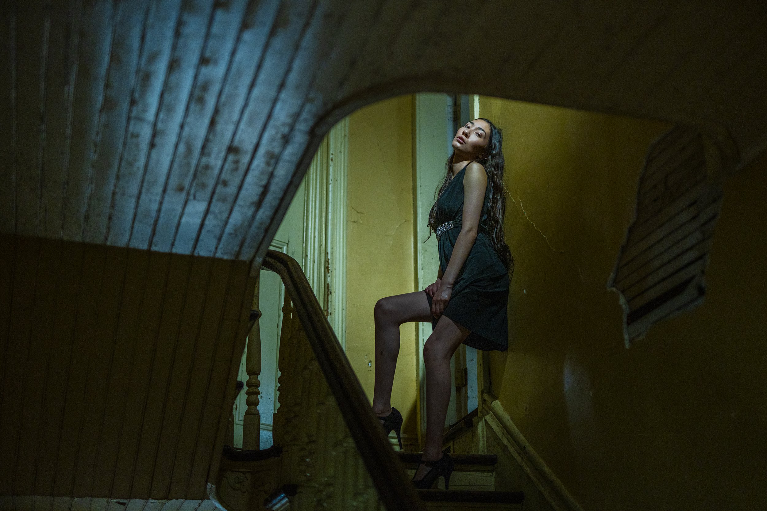 A woman with long dark hair in a black dress standing on a staircase, seen through a decorative opening in a wooden structure, with yellow and green walls behind her.
