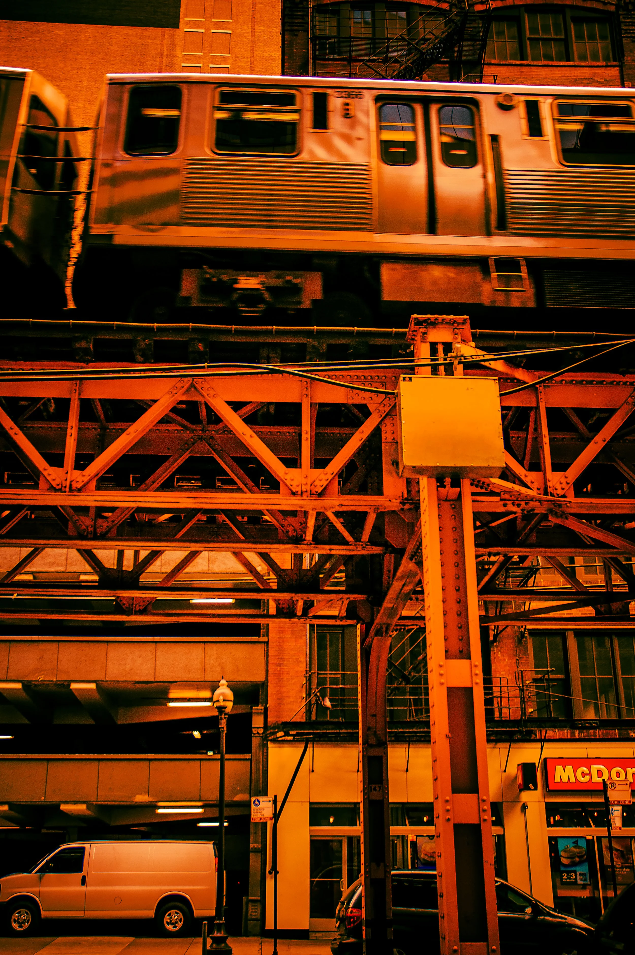 A city scene at night with a moving train on an elevated track, brick buildings in the background, and a McDonald's sign below.