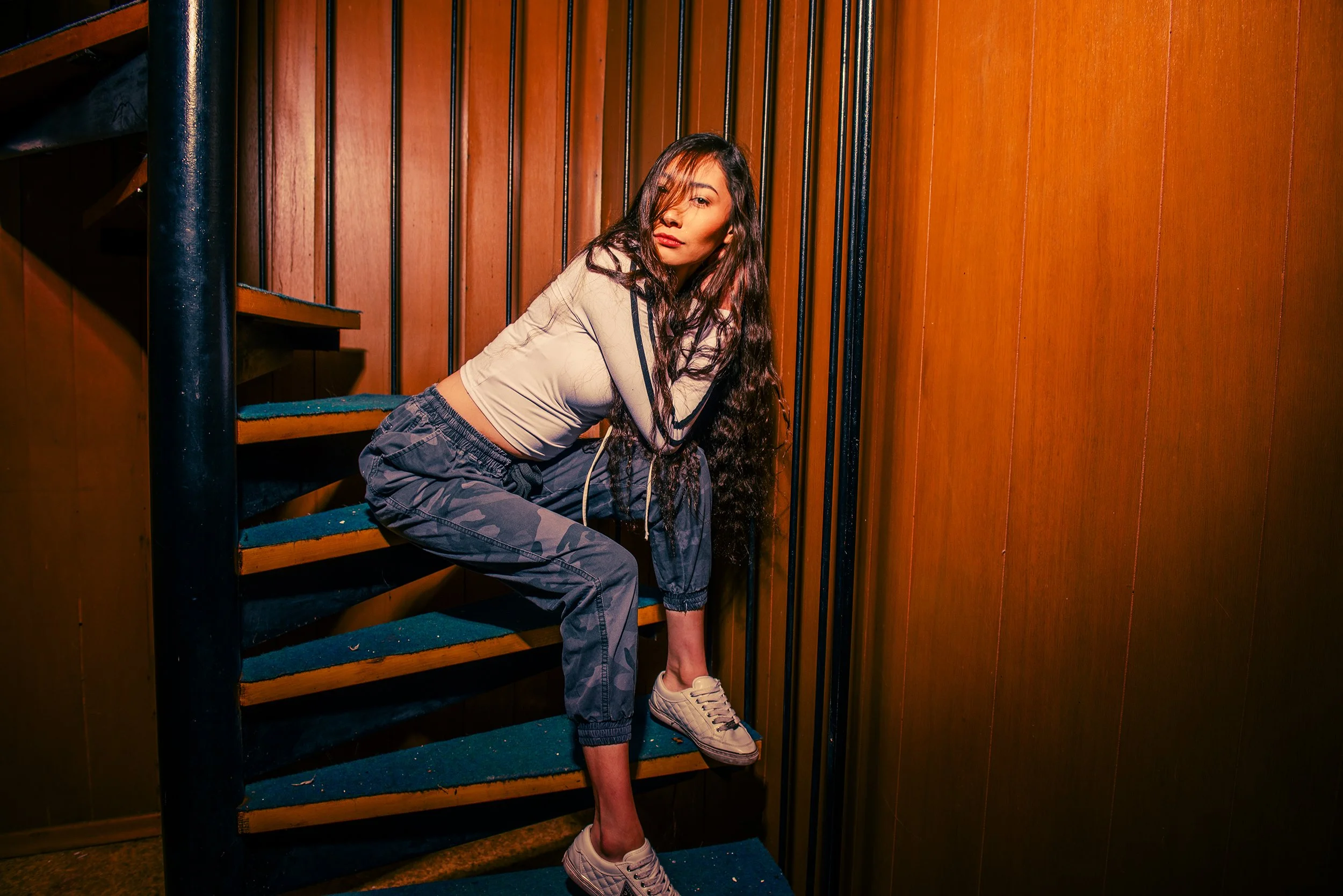 A young woman with long dark curly hair sitting on a staircase against a wooden-paneled wall. She is wearing a beige long-sleeve crop top, camouflage pants, and white sneakers, with a relaxed and confident expression.