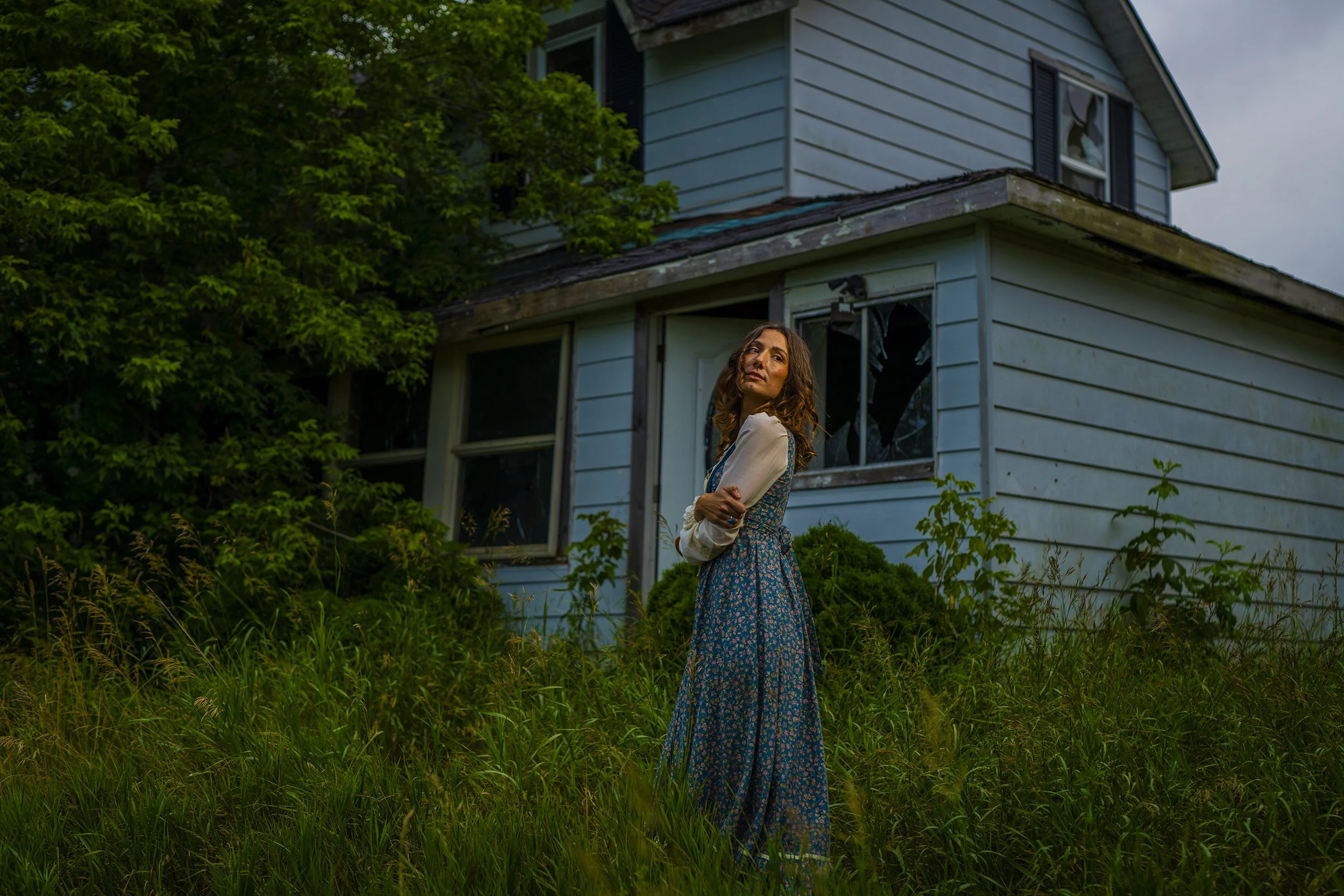A woman in a long floral dress stands outside a blue wooden house with broken windows, surrounded by overgrown grass and trees.