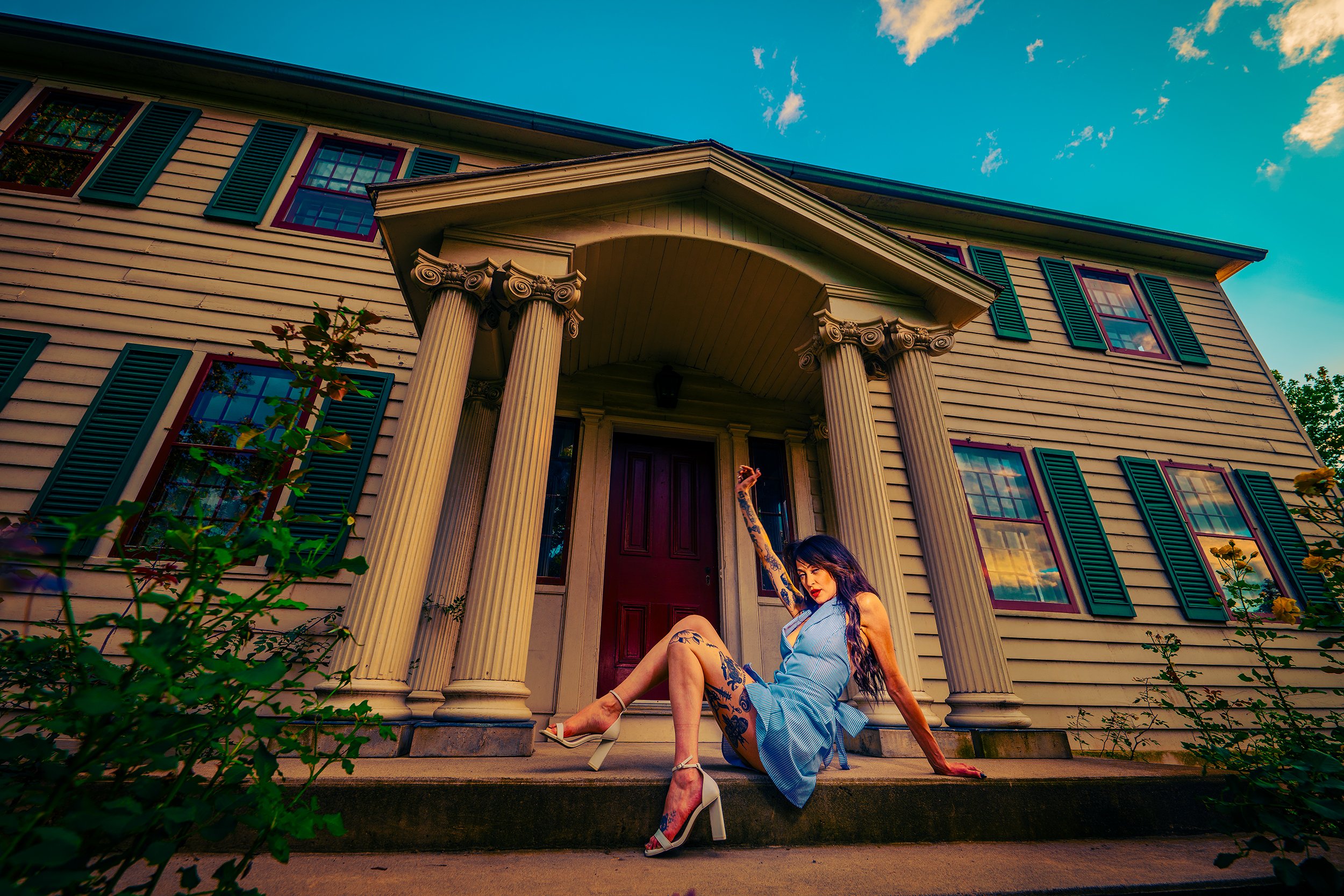 Woman in a light blue dress with tattoos sitting on the front porch of a yellow house with green shutters, columns, and a red door, during sunset.