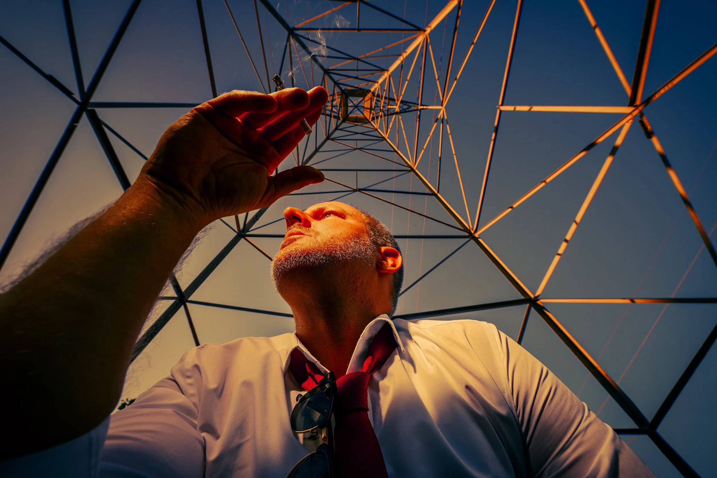 A man in a white shirt and red tie looking up at an orange power transmission tower during sunset, with the sky in the background.
