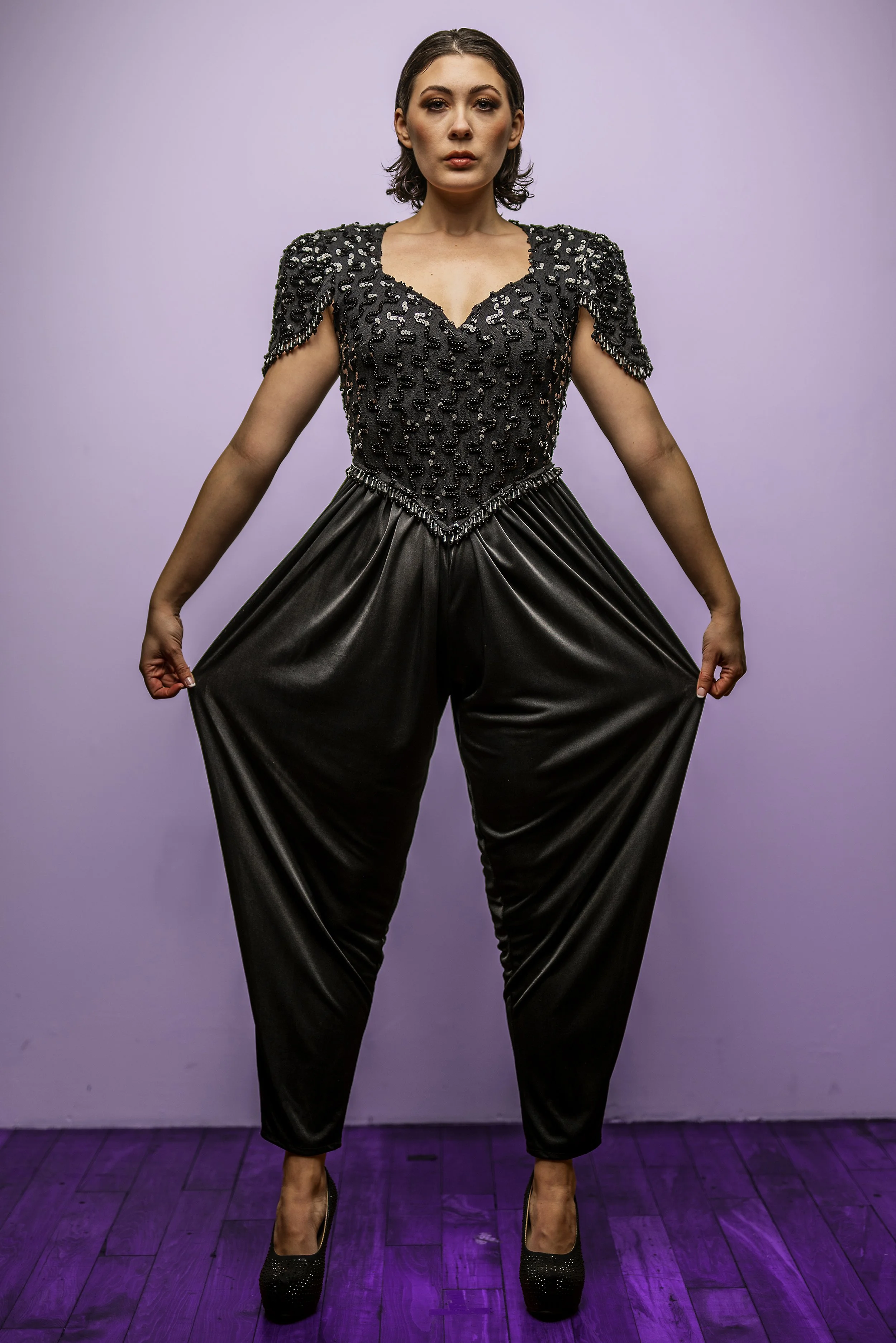 A woman with light skin and short dark hair standing against a purple wall, wearing a black embellished top, black satin pants, and black high heels, with a confident expression and stance.