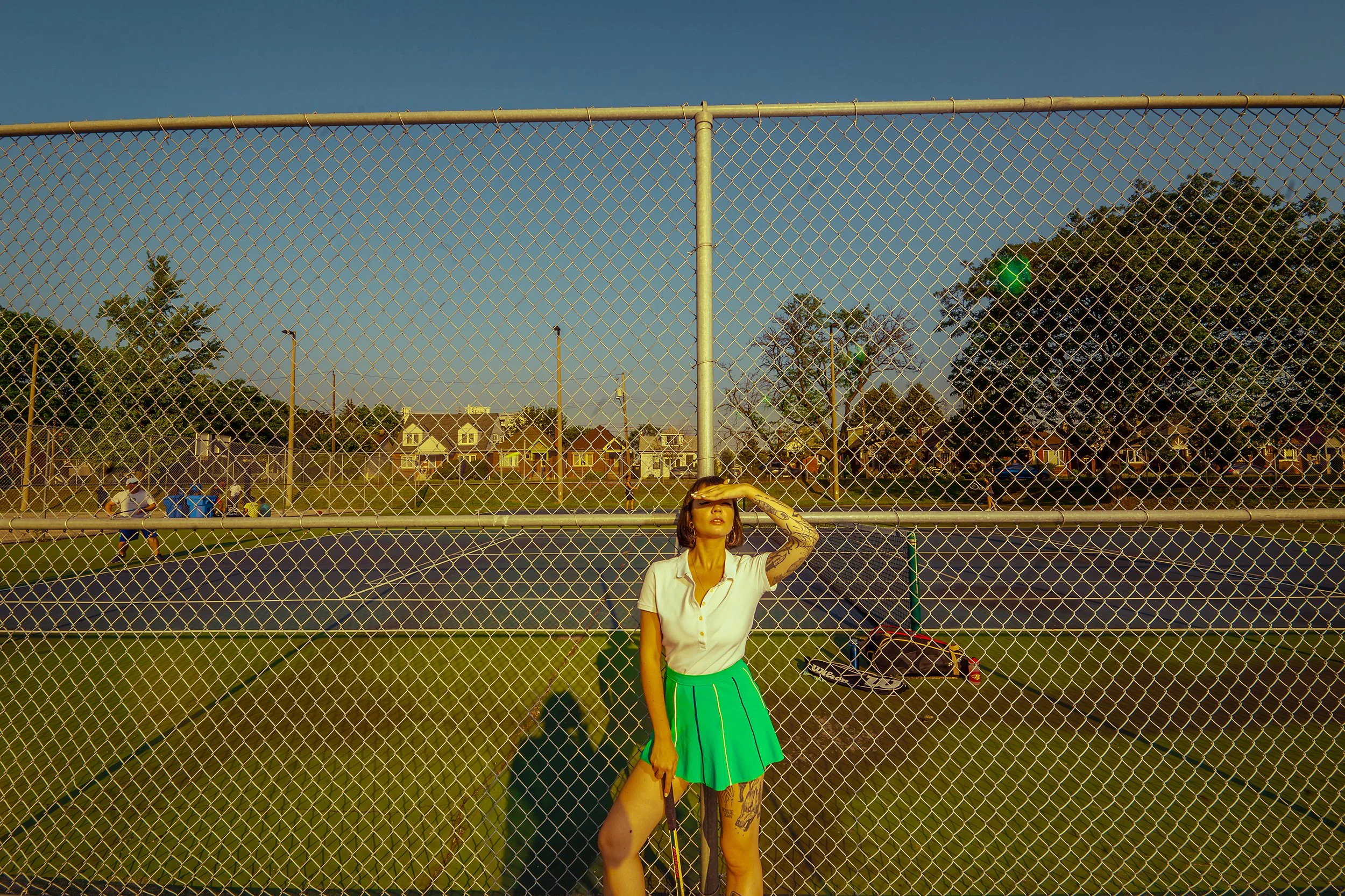 A woman with tattoos on her left arm, standing behind a chain-link fence on a tennis court, shielding her eyes from the sun with her hand, wearing a white shirt and a bright green skirt, with a sunny sky and houses in the background.