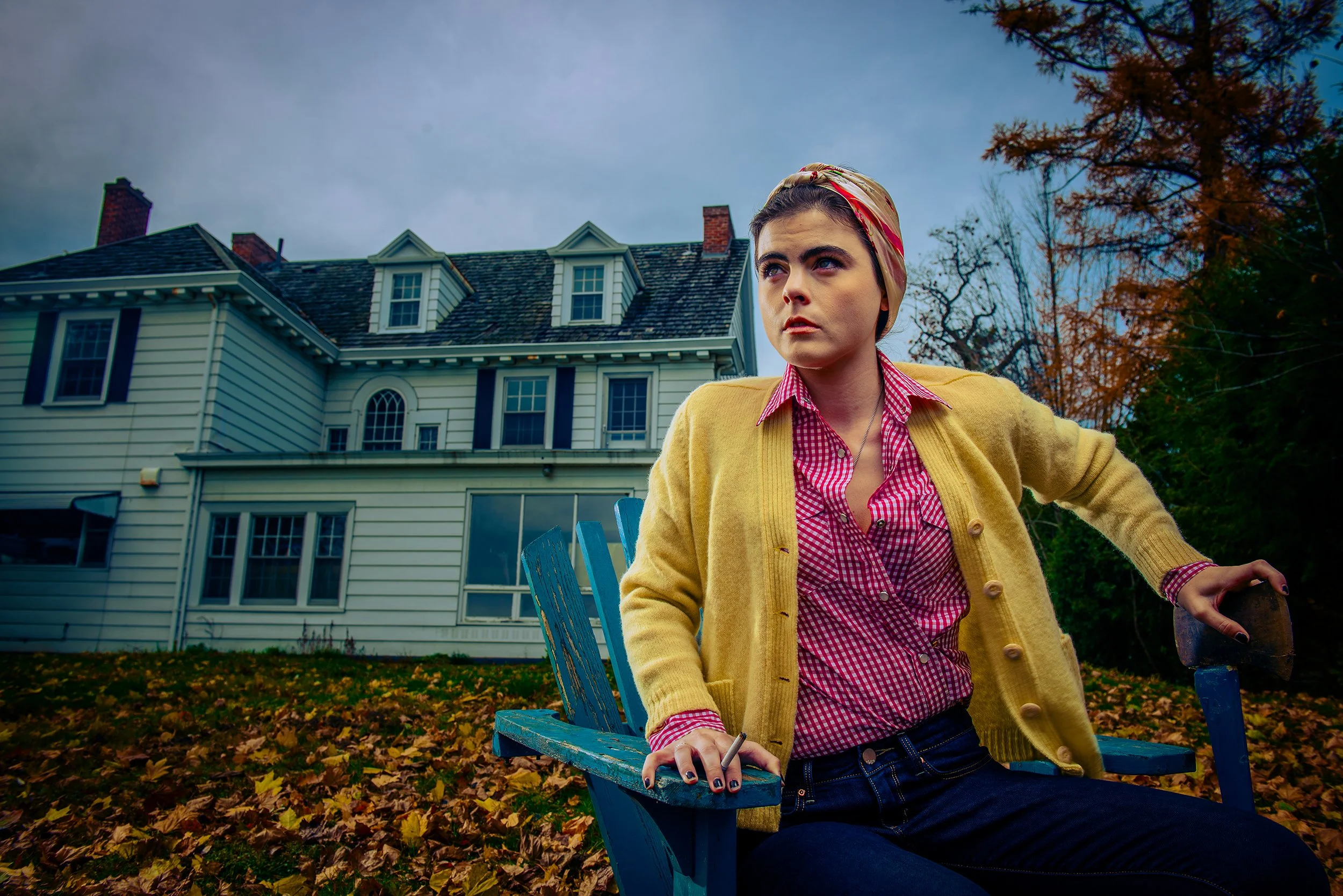 A woman with dark hair and blue eyes wearing a pink checkered shirt, yellow cardigan, and headscarf, sitting on a blue wooden bench outside in front of a large house with white siding and multiple chimneys, surrounded by autumn leaves and trees with 