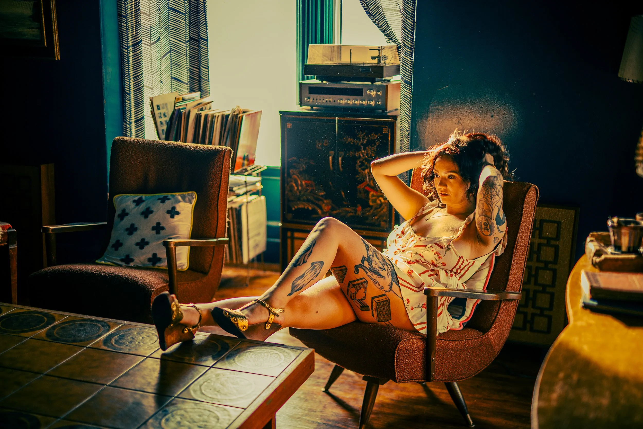 Woman with tattoos sitting in a vintage armchair in a cozy, dimly lit room with sunlit window, patterned curtains, and record player in the background.