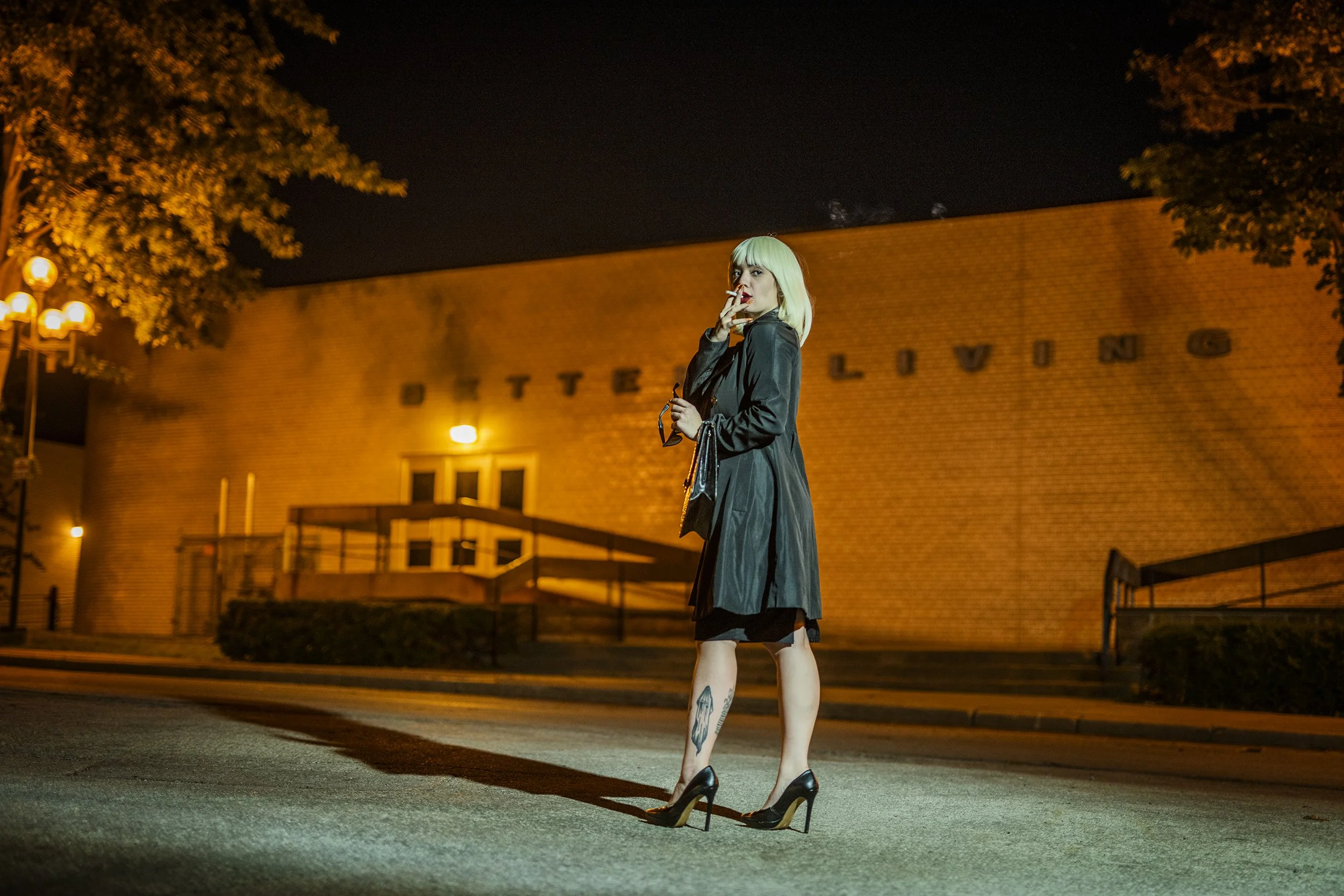 A woman with platinum blonde hair wearing a black trench coat and high heels standing on a lit street at night in front of a brick church with the words 'BETHEL V' visible on the building.