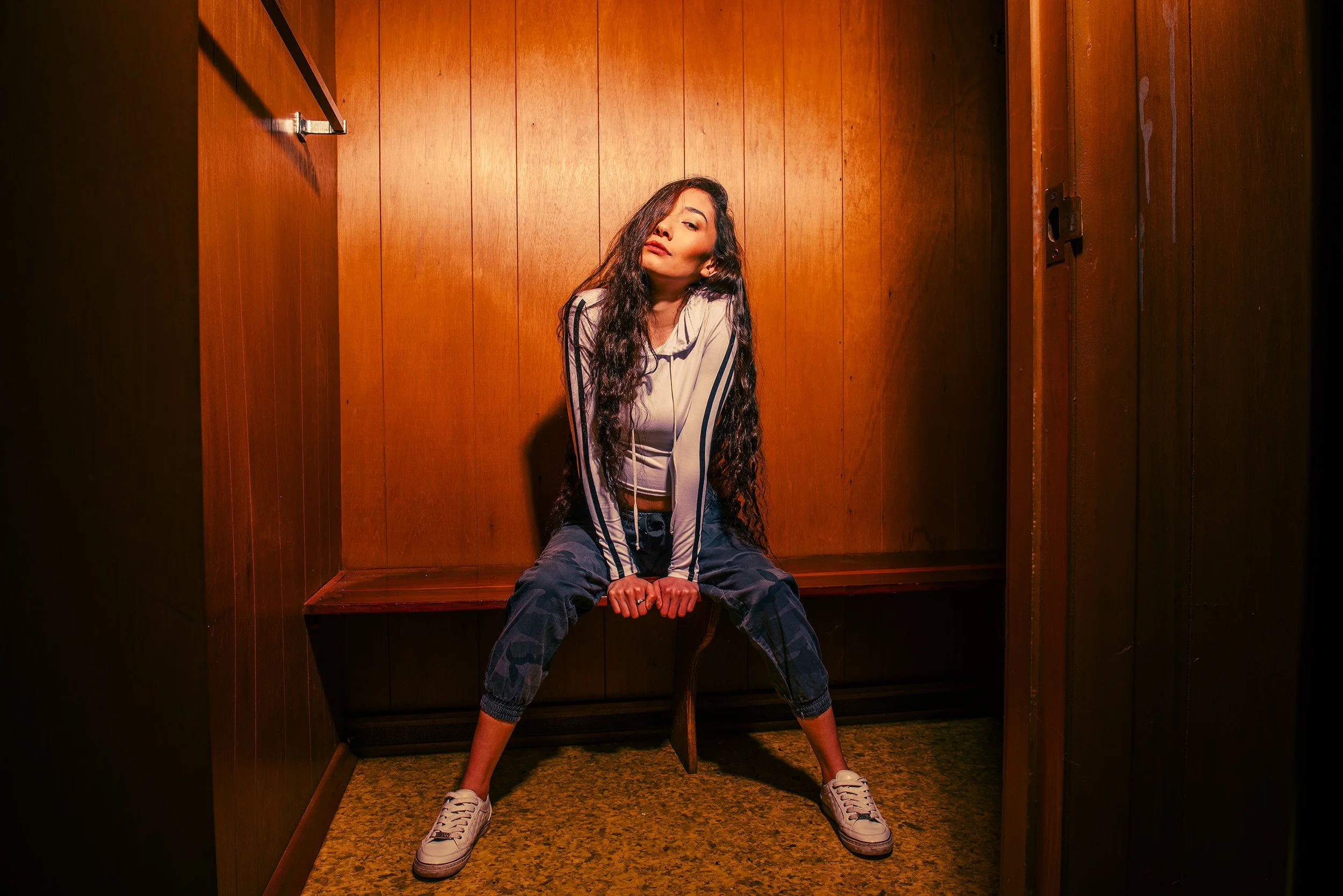 Woman with long curly hair sitting inside a wooden box or elevator with walls and floor made of wood, wearing a light jacket, camo pants, and white sneakers, with a confident expression.