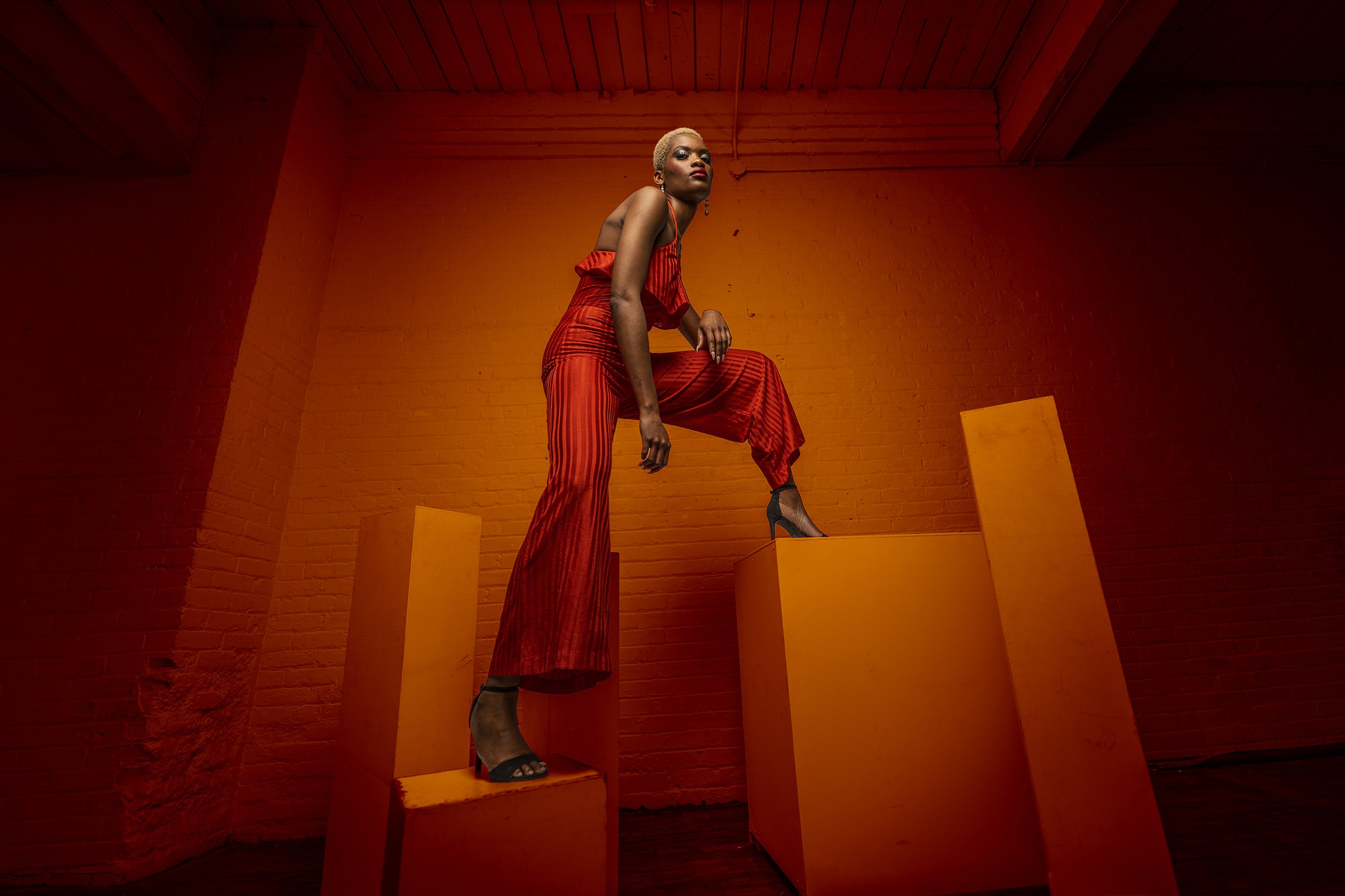A woman with short blonde hair wearing a red striped dress and black high heels is standing on a yellow pedestal in a room with orange brick walls and ceiling beams. The lighting casts red and orange hues.