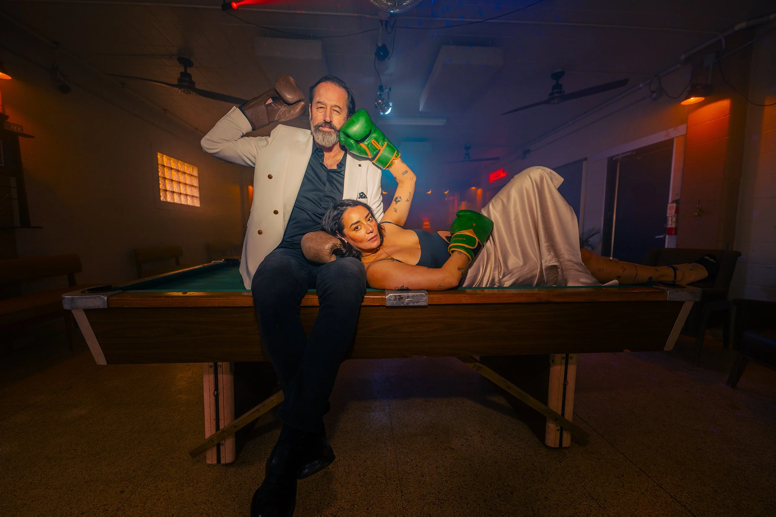 A man and woman posing on a pool table in a dimly lit room. The man is standing behind the woman, holding a boxing glove to his ear like a phone, and the woman is lying on the table wearing boxing gloves, looking at the camera.